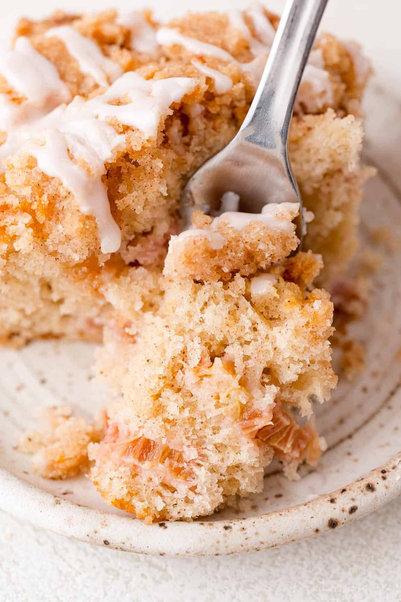A close-up of a fork cutting into a slice of crumbly, iced coffee cake on a white ceramic plate.