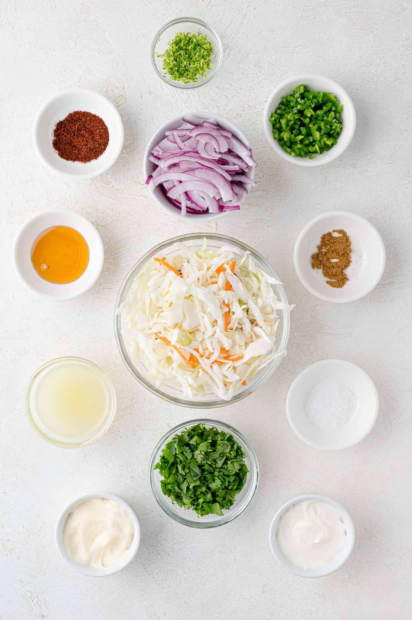 A variety of ingredients for taco slaw arranged in small bowls on a white surface, including cabbage, carrots, red onion, herbs, spices, mayonnaise, lime juice, and sour cream.