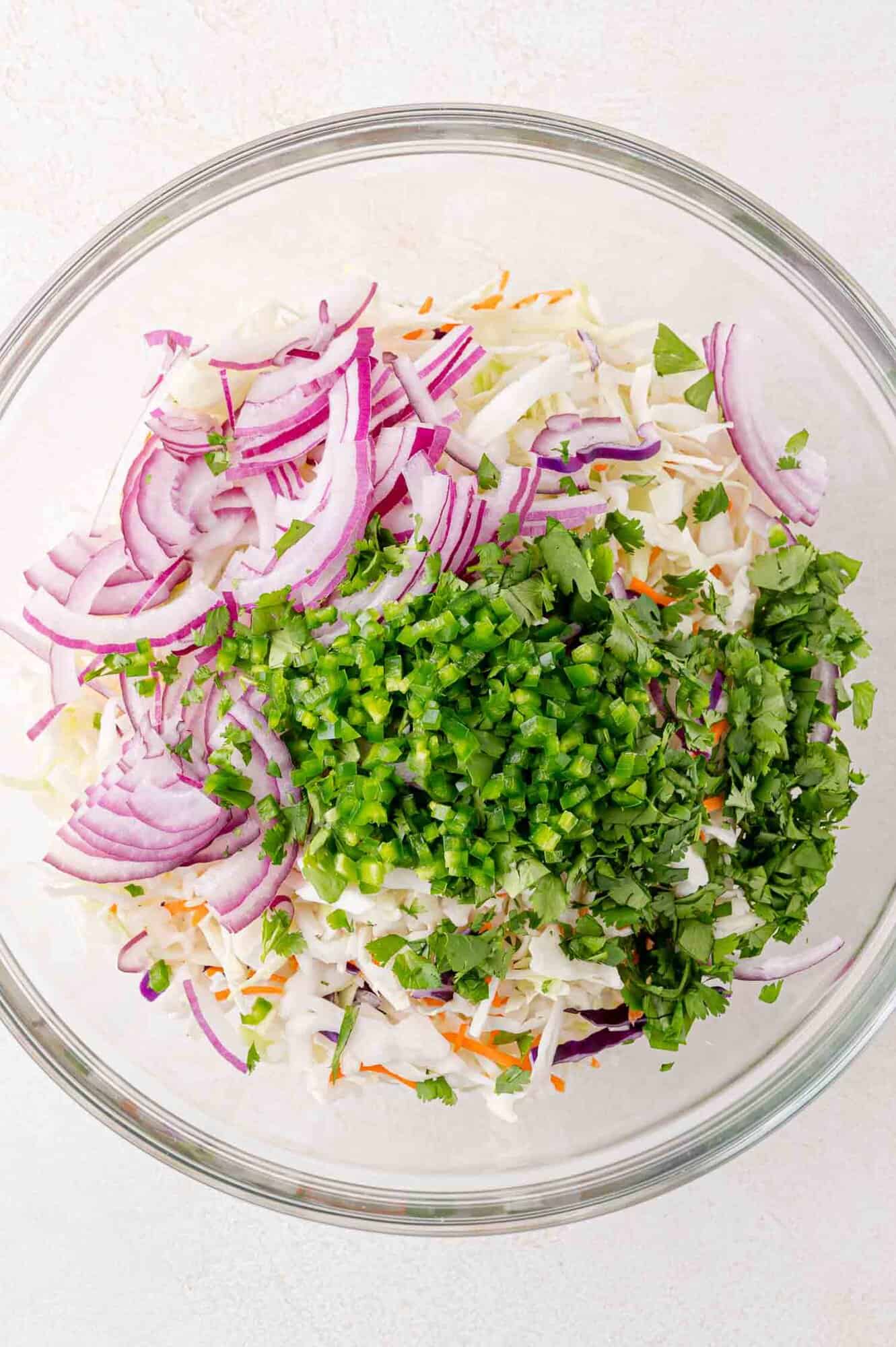 A glass bowl filled with sliced red onions, chopped green herbs, diced jalapeños, shredded cabbage, and carrots on a light surface.