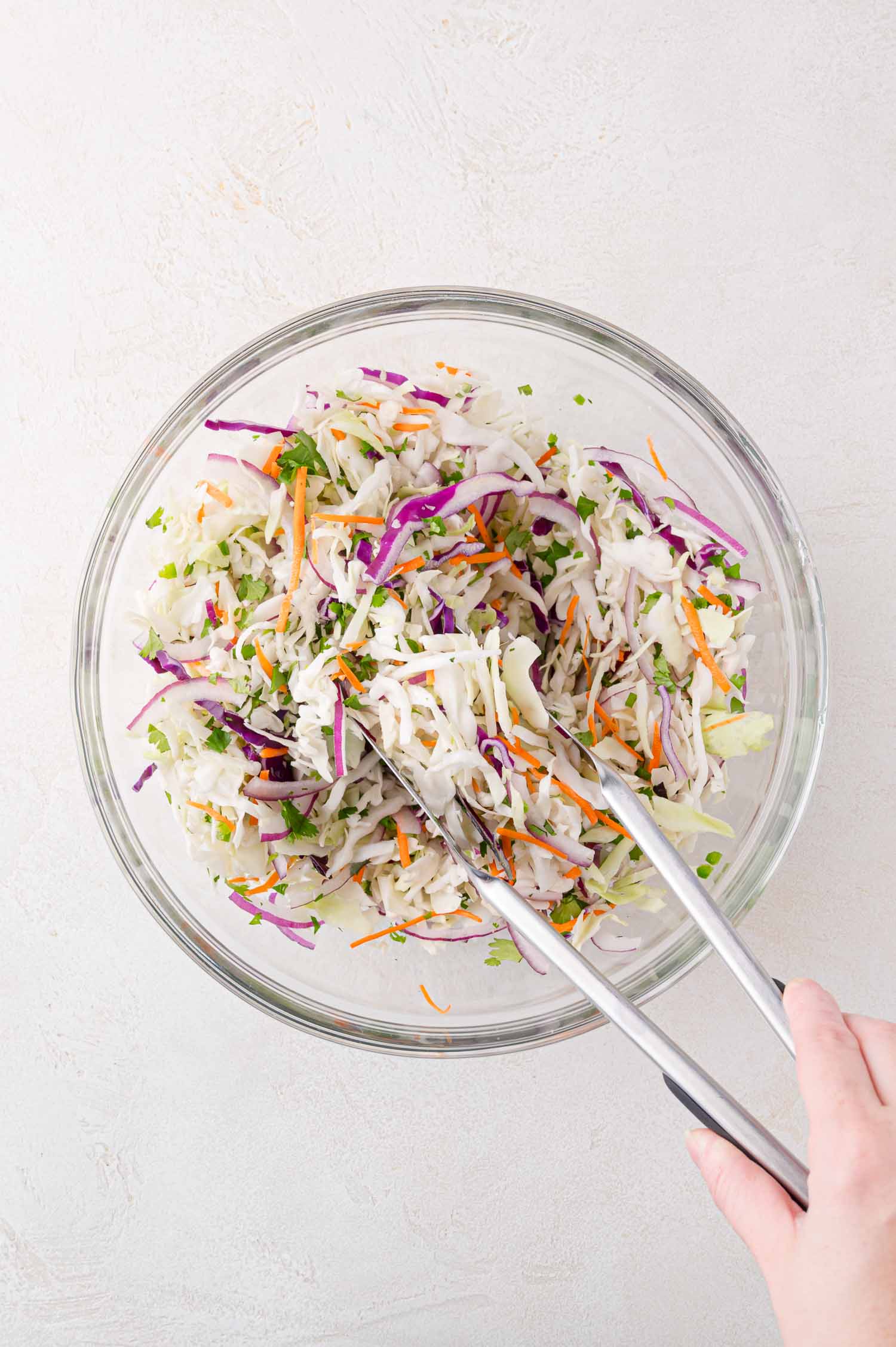 A hand uses metal tongs to mix shredded cabbage, carrots, and herbs in a large clear glass bowl on a light surface.