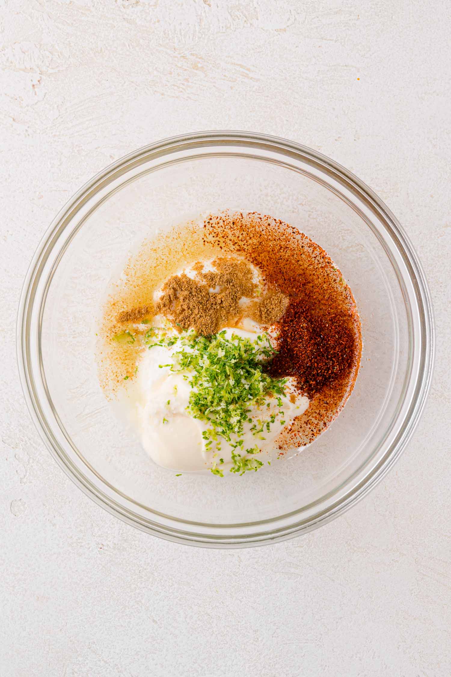 A glass bowl containing white sauce topped with green herbs, brown spices, and red seasoning, placed on a light-colored surface.