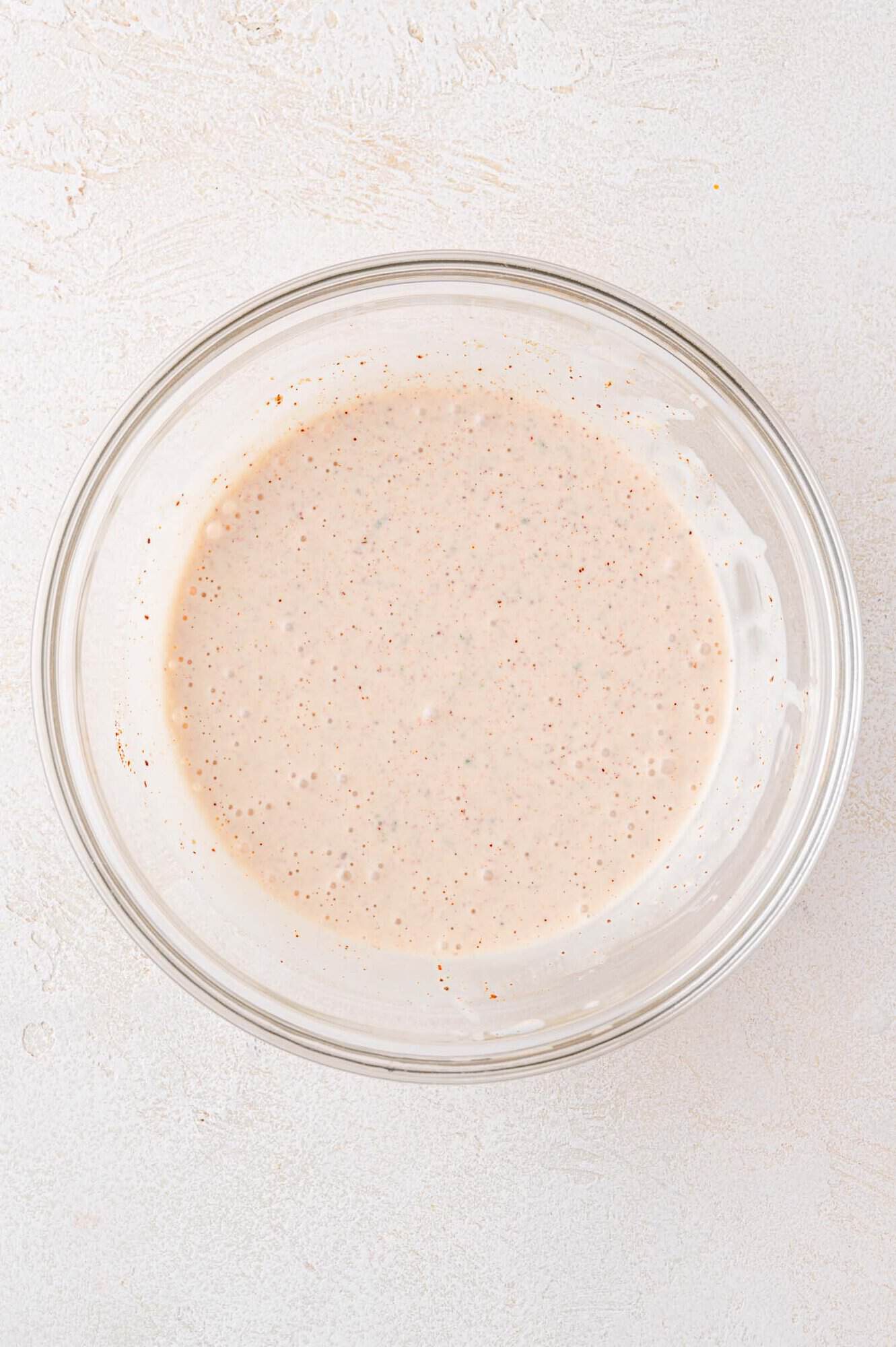 A glass bowl filled with a creamy, speckled sauce sits on a light-colored textured surface.