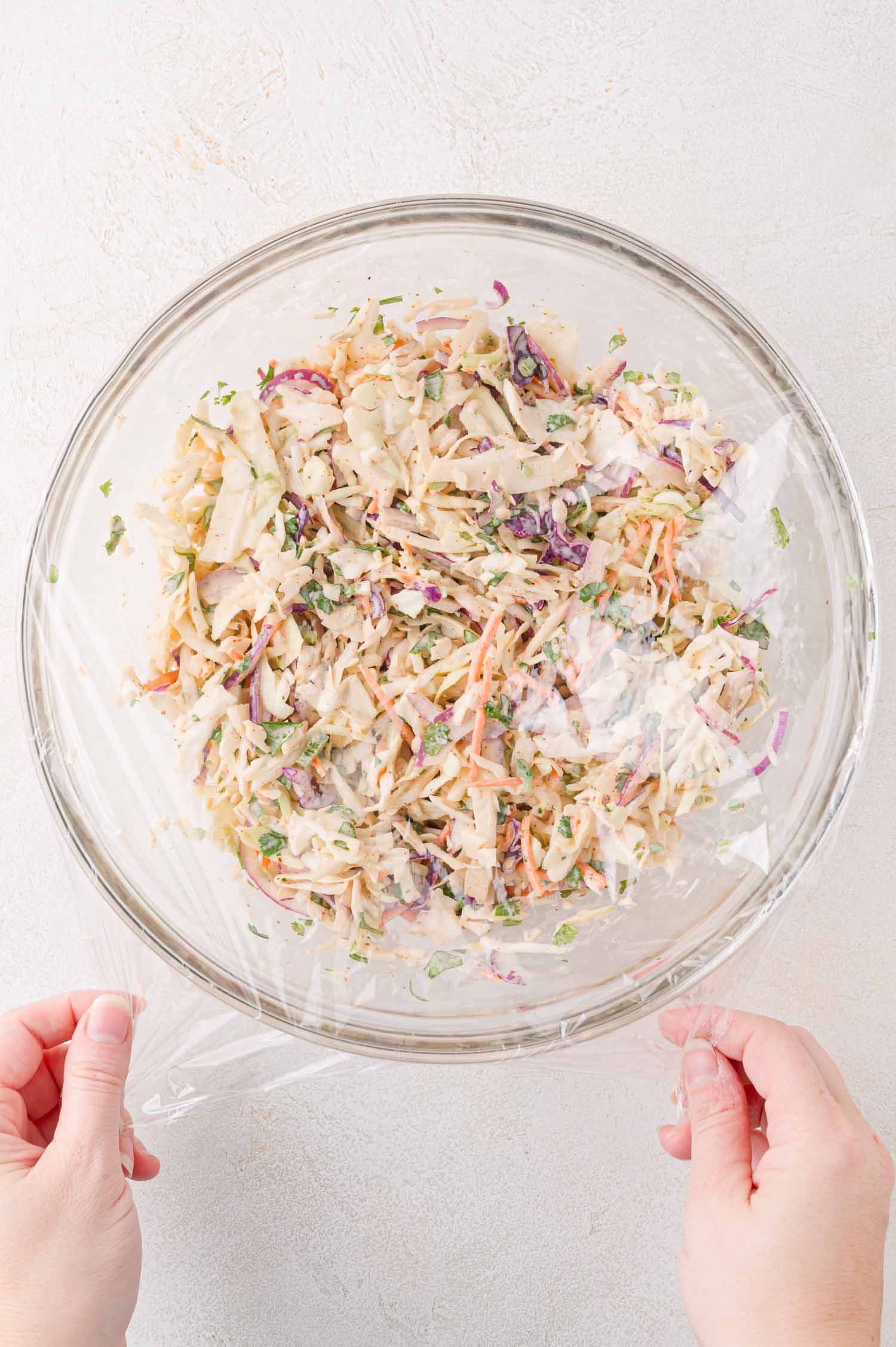 Hands covering a glass bowl of coleslaw with plastic wrap on a light-colored surface.
