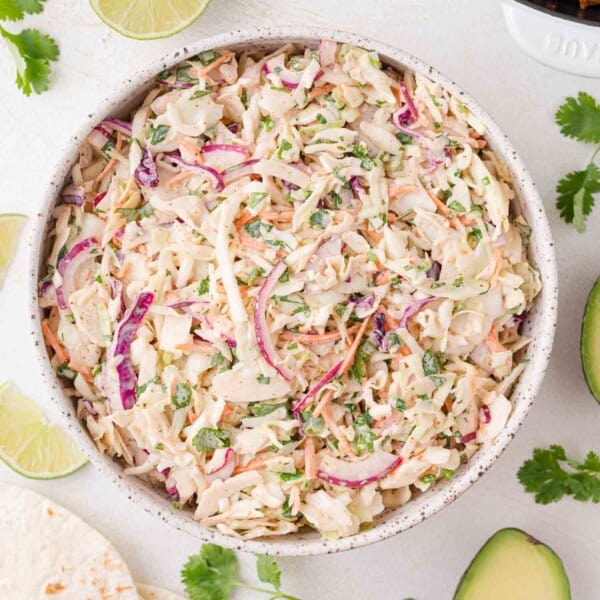 A bowl of creamy taco slaw with shredded cabbage, carrots, red onion, and chopped herbs, surrounded by lime wedges, avocado, and cilantro.
