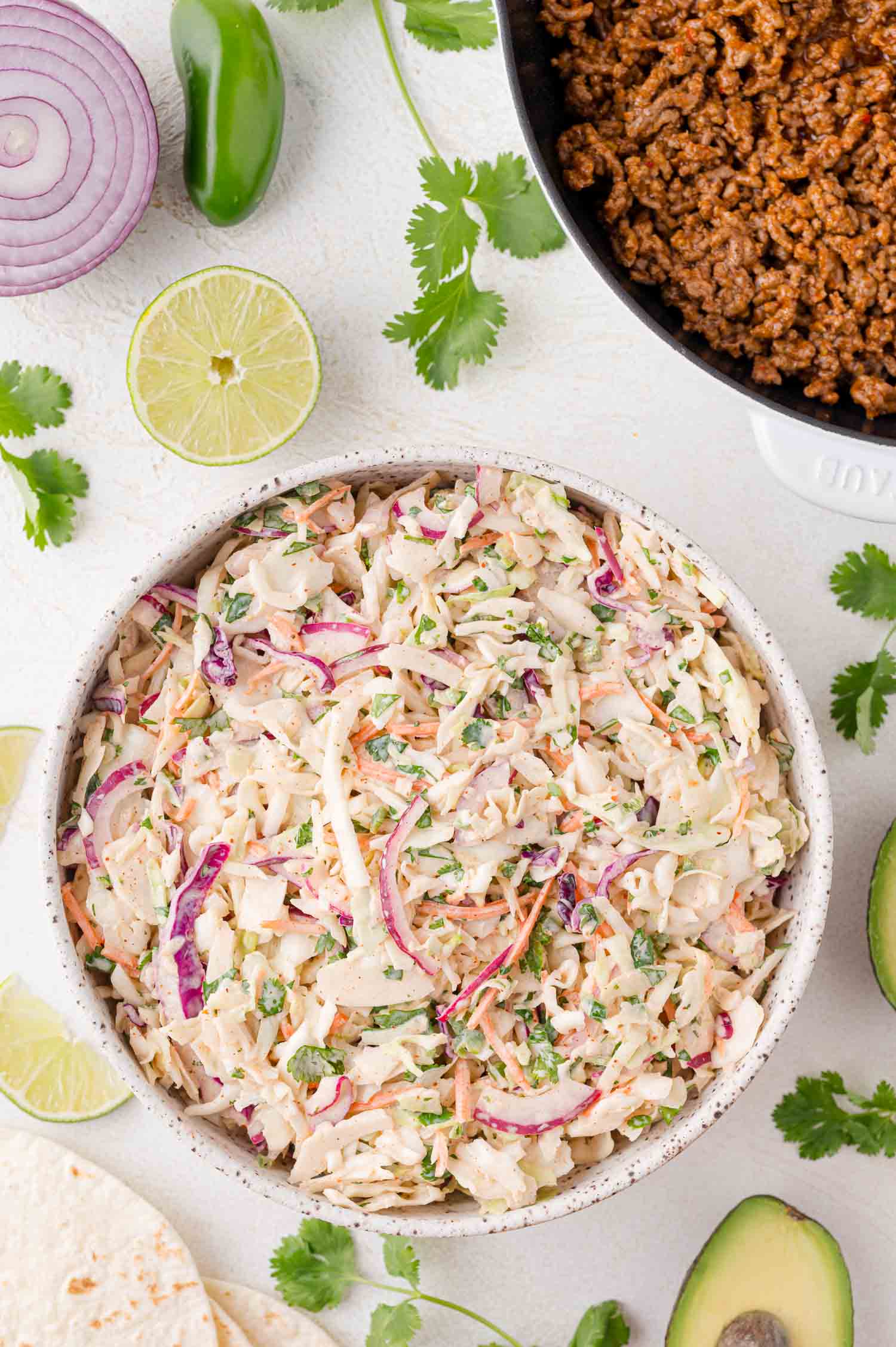 A bowl of creamy taco slaw with red onions and cilantro, surrounded by sliced lime, avocado, red onion, jalapeño, ground meat, and tortillas on a white surface.