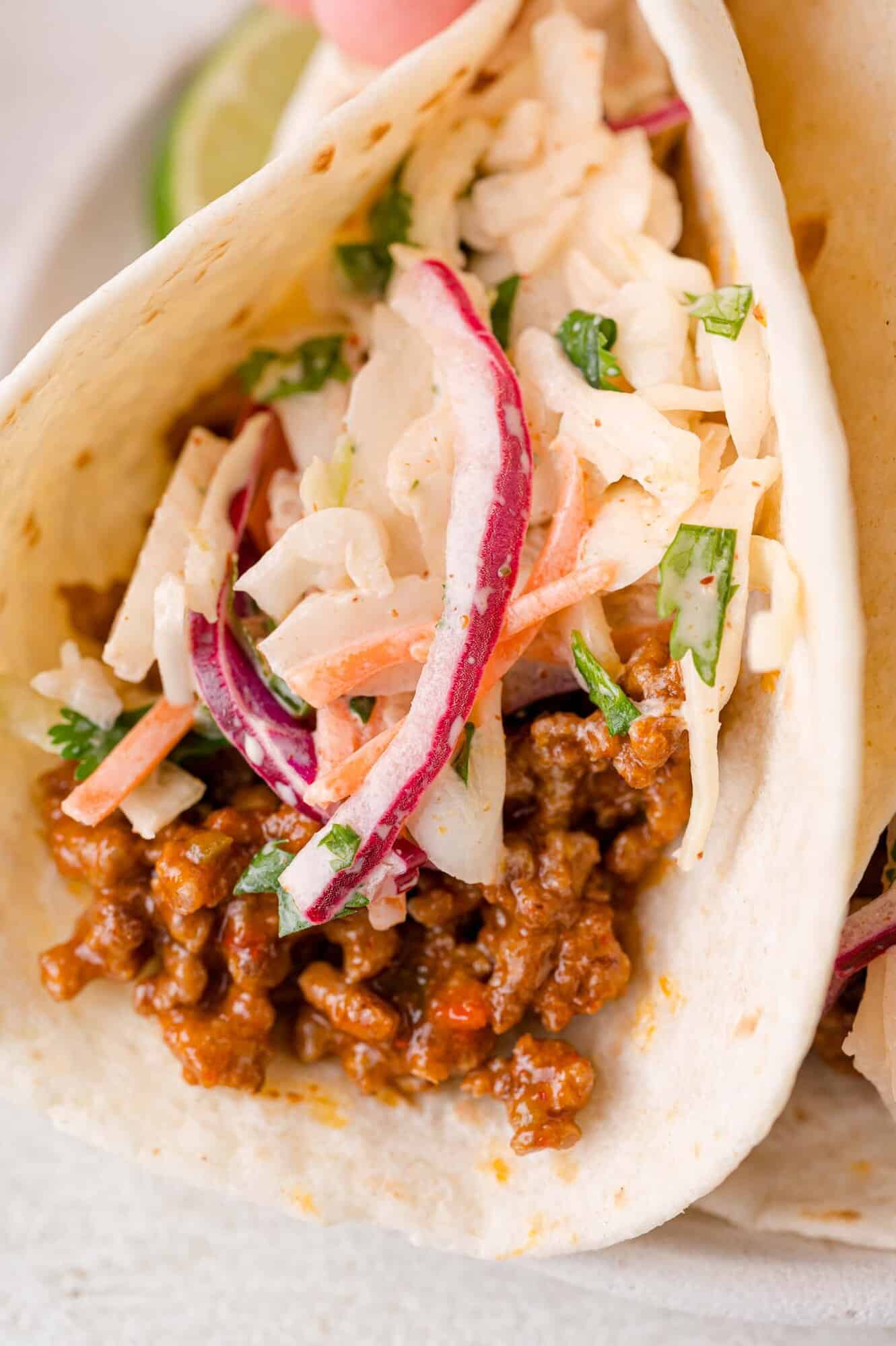A soft taco filled with seasoned ground beef and topped with a slaw made of cabbage, red onion, carrots, and herbs.