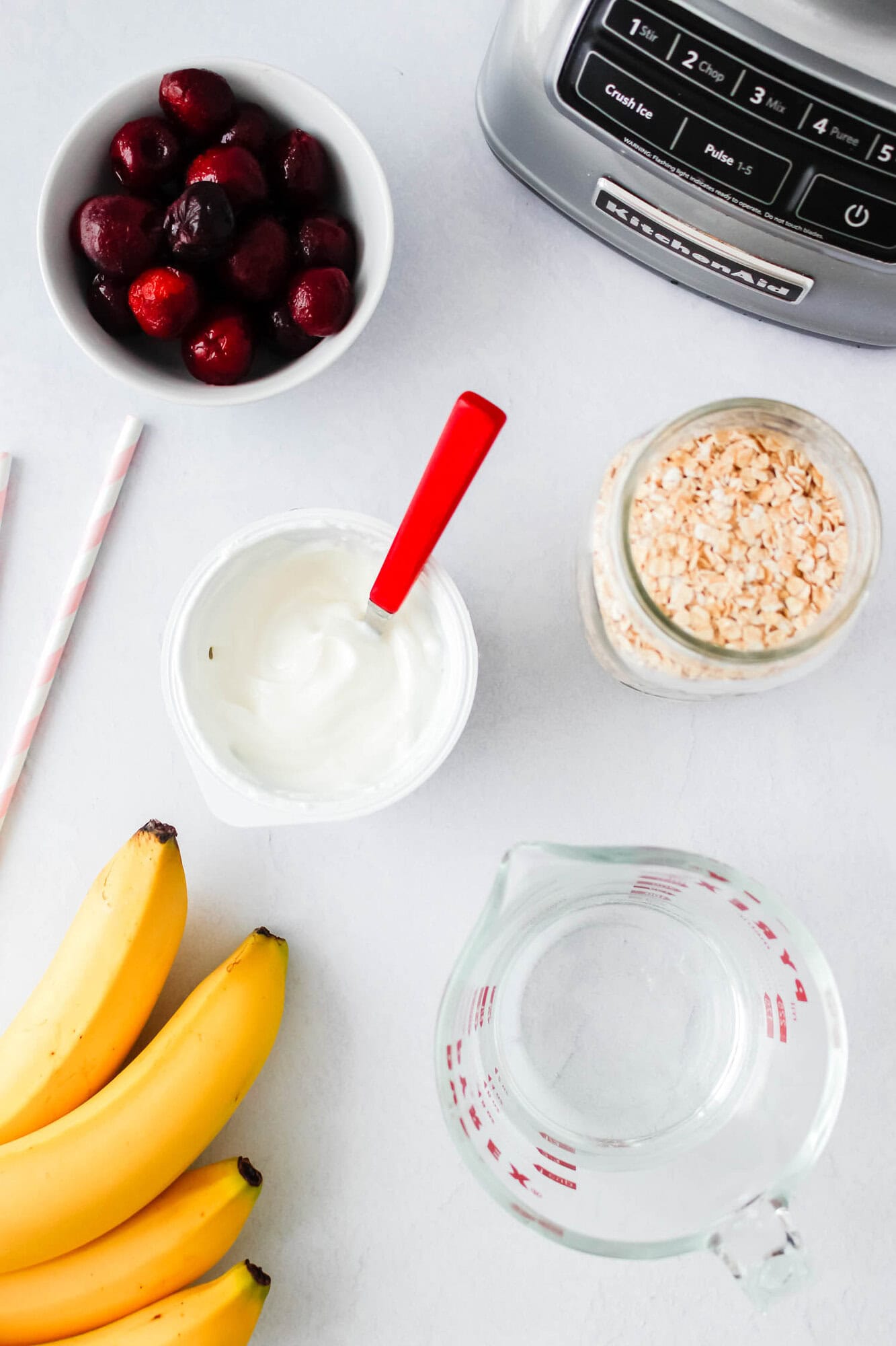 A countertop with bananas, a bowl of cherries, a bowl of oats, a container of yogurt with a red spoon, a glass measuring cup, two straws, and part of a blender visible.