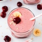 A glass of pink smoothie topped with banana slices, a cherry, and oats, with a striped straw and scattered cherries and banana slices nearby.