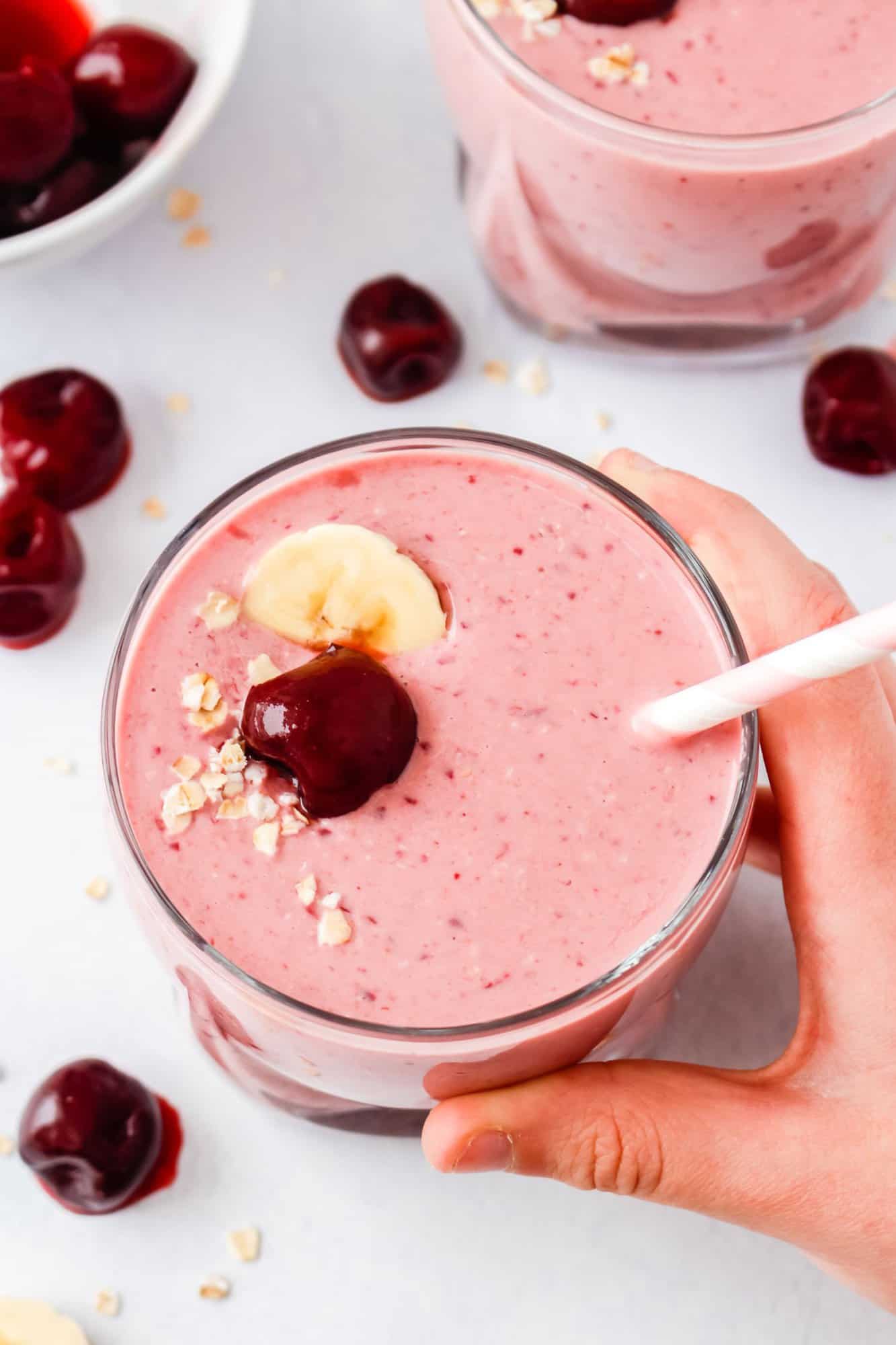 Cherry Smoothie 3 A hand holds a glass of pink cherry smoothie topped with a banana slice, cherry, and oats, with a straw inserted. Other cherries are scattered on the white surface.