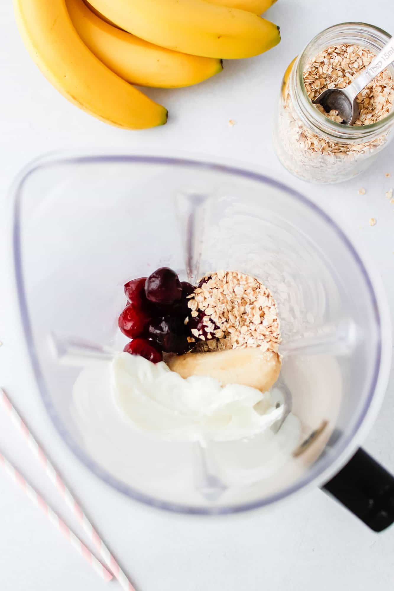 A blender contains yogurt, oats, cherries, and a banana. Nearby are bananas, a jar of oats with a spoon, and striped straws on a white surface.