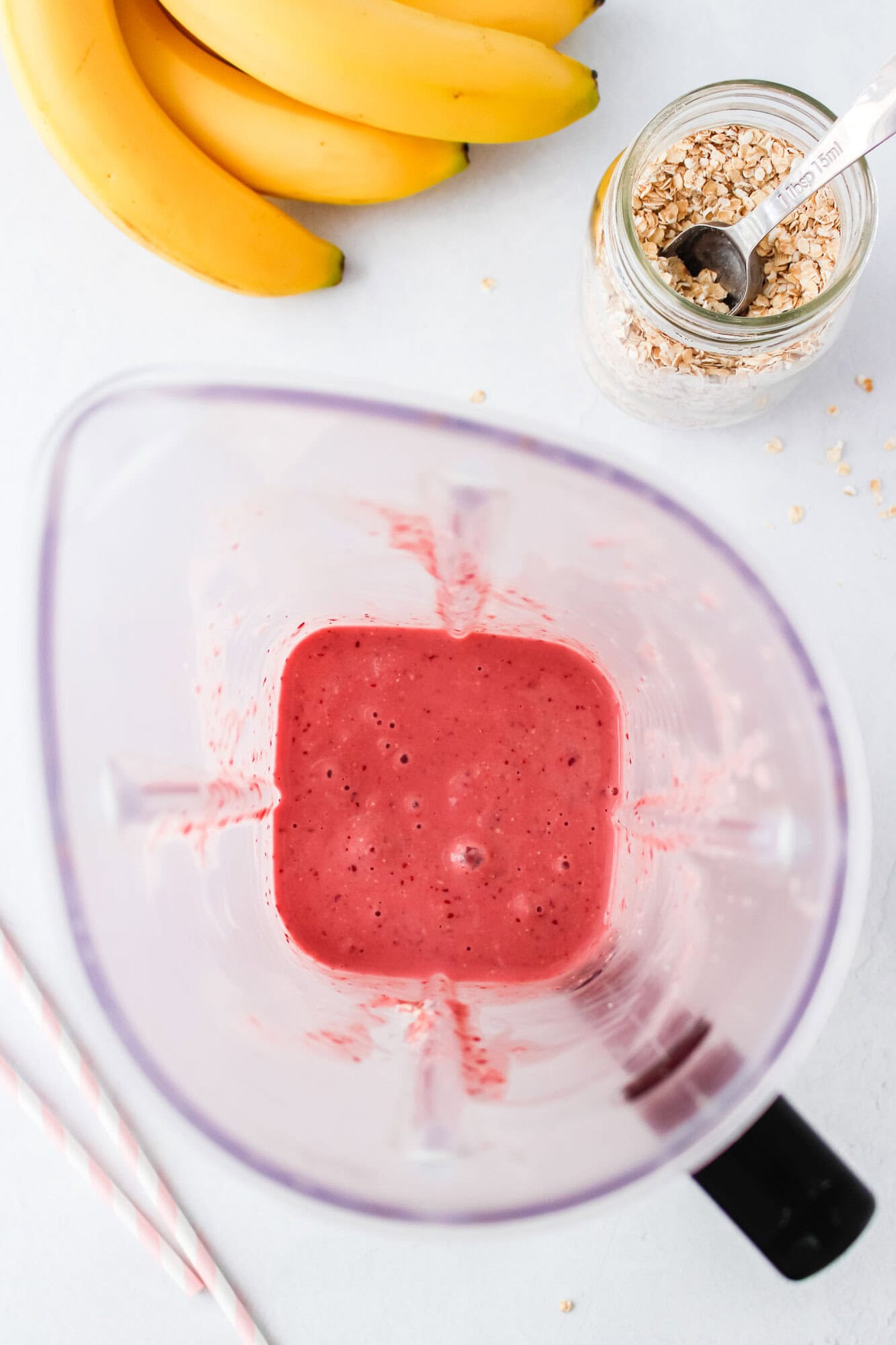 A blender containing pink smoothie sits on a white surface, with bananas, a jar of oats with a spoon, and two striped straws nearby.