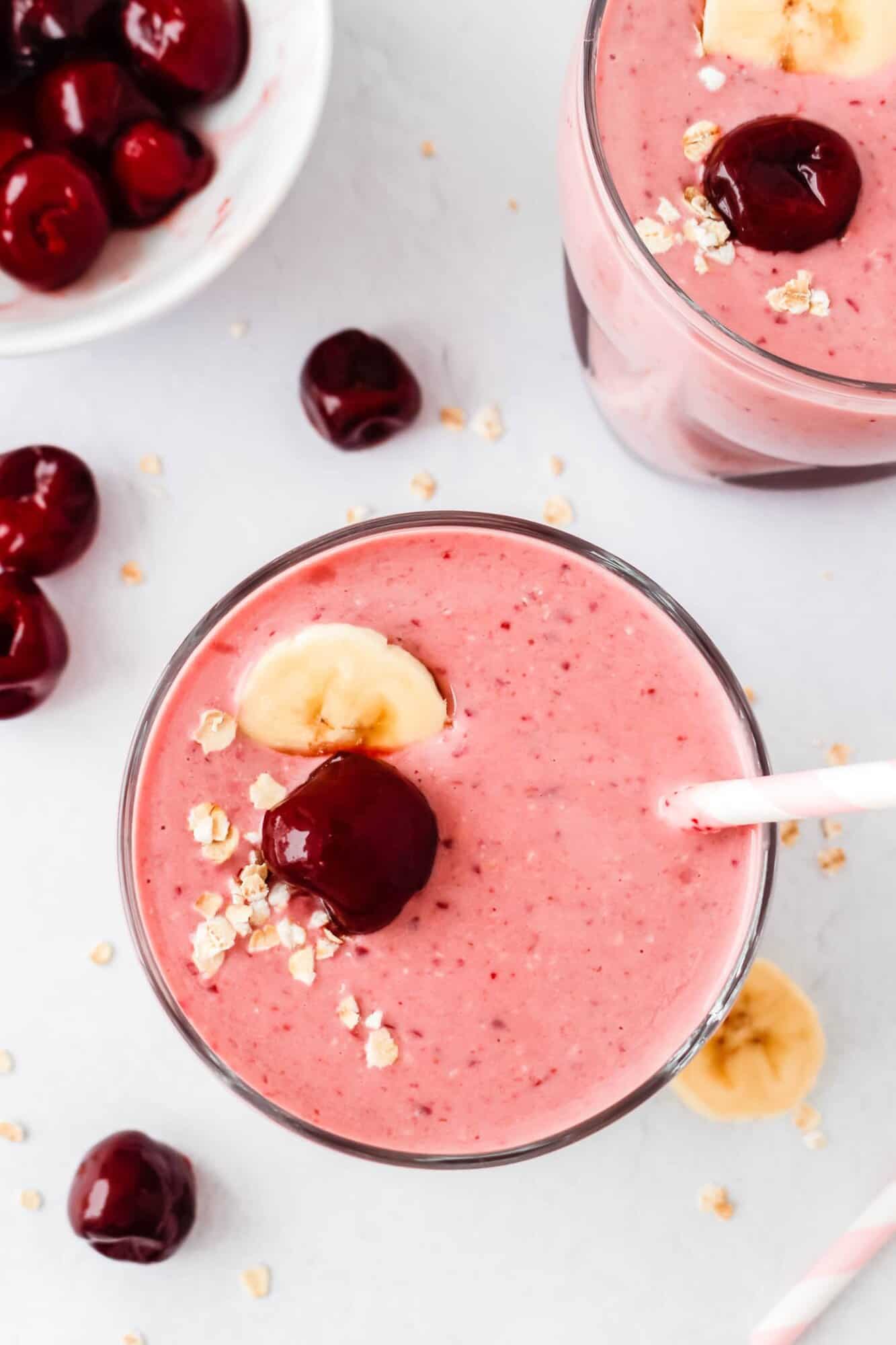 Cherry Smoothie 2 A glass of pink smoothie topped with a cherry, banana slices, and oats, with a straw; whole cherries and oats are scattered on the table nearby.