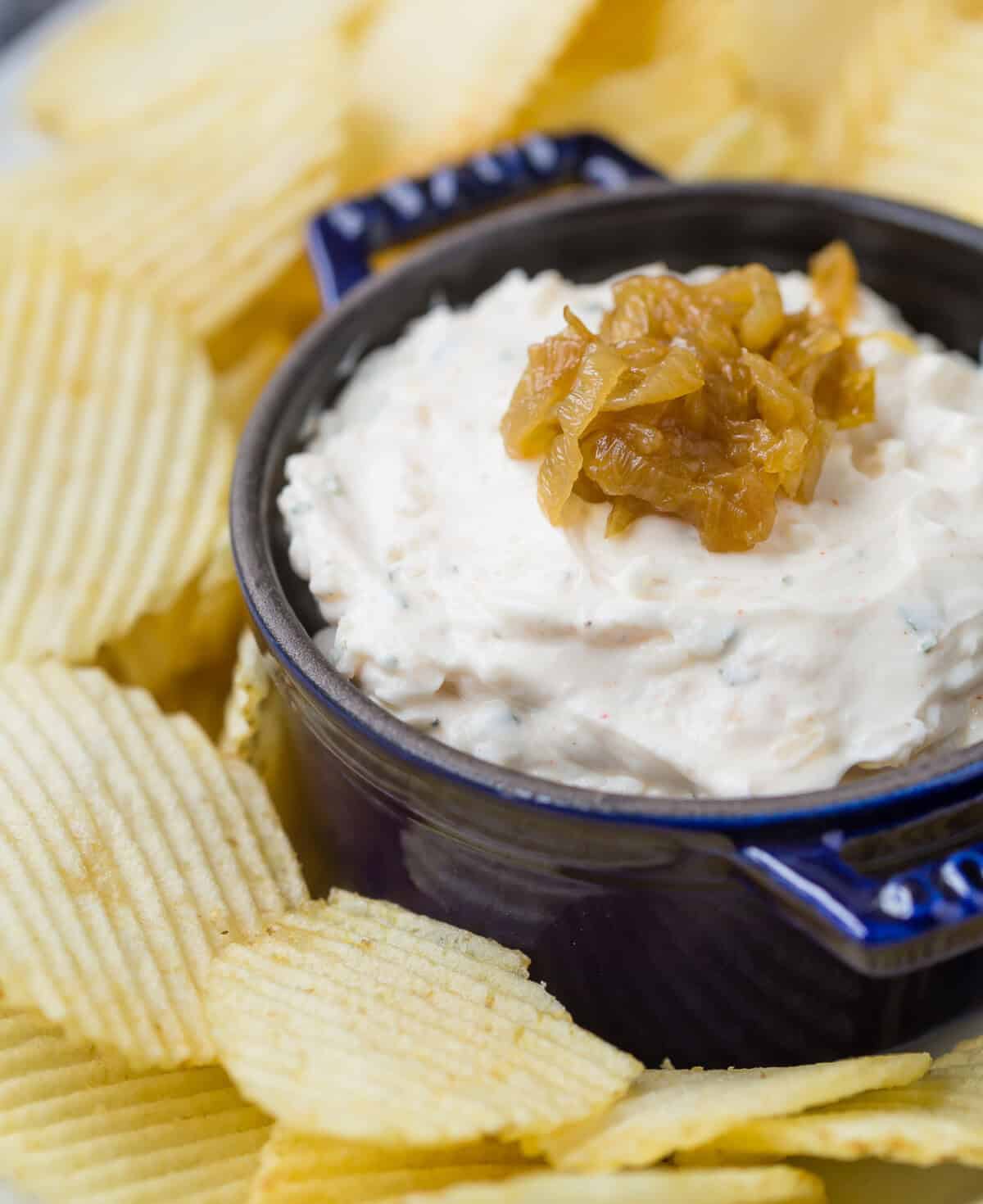 A dark blue bowl of creamy french onion dip dip topped with caramelized onions is surrounded by ridged potato chips.