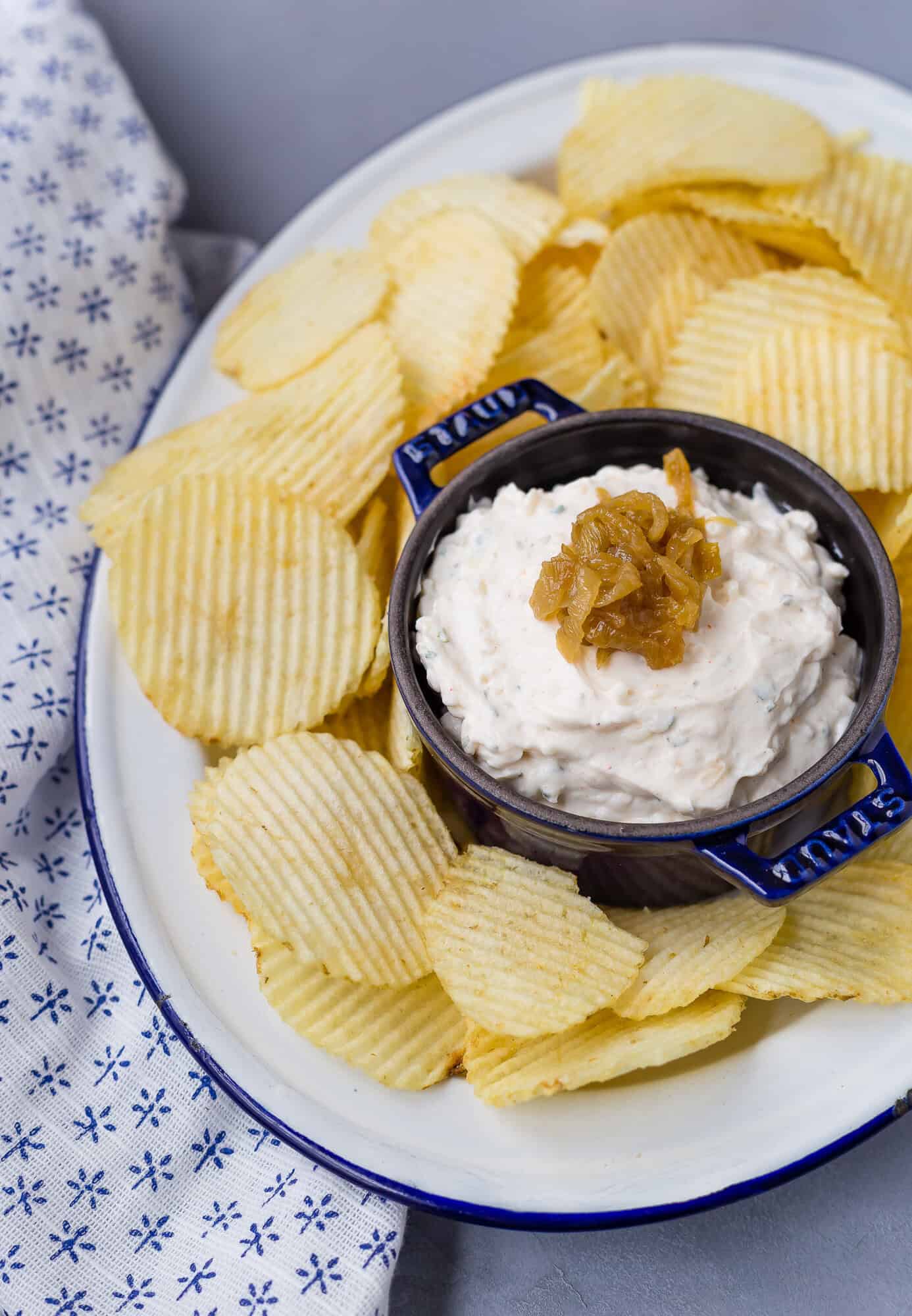 A plate of ridged potato chips surrounds a bowl of creamy dip topped with caramelized onions, next to a patterned cloth.