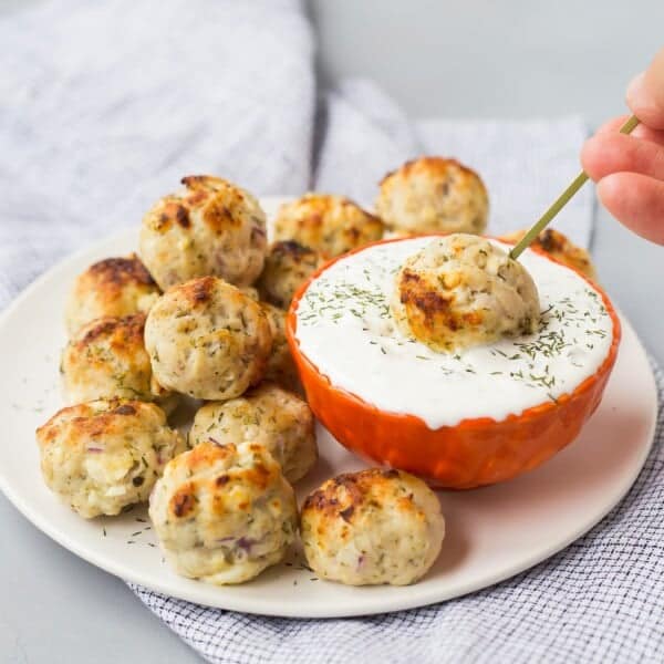 A plate of baked Greek turkey meatballs next to a bowl of white dipping sauce, with one meatball being dipped using a skewer.