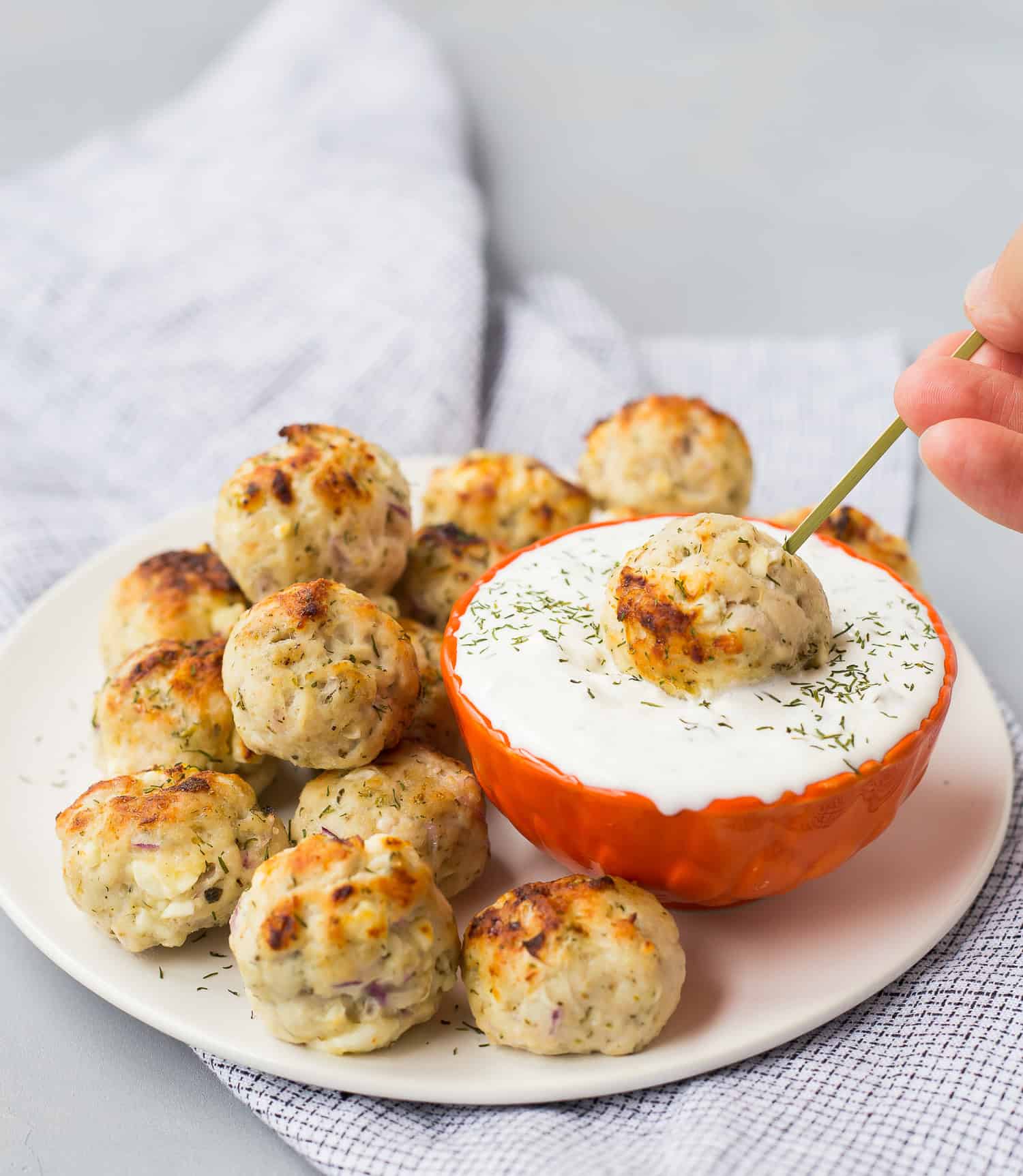 A plate of baked  greek turkey meatballs is served with a bowl of white dipping sauce; one meatball is being dipped into the sauce using a skewer.