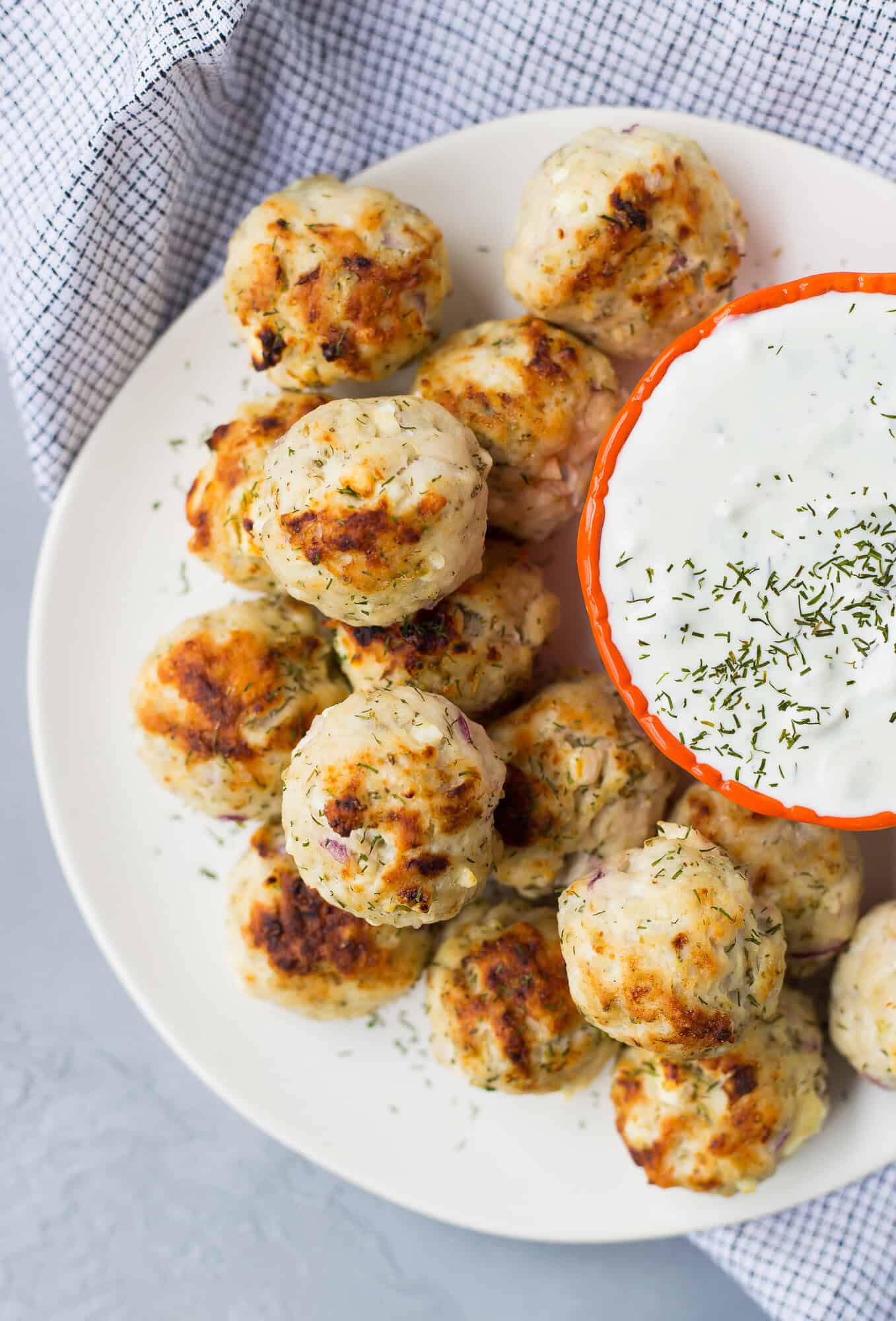 A white plate with several Greek turkey meatballs next to a small bowl of creamy dipping sauce, sprinkled with herbs, on a gray surface with a checkered cloth.