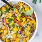 A bowl of mango slaw with cabbage, carrots, red onion, cilantro, and lime wedges, mixed with a black serving spoon.