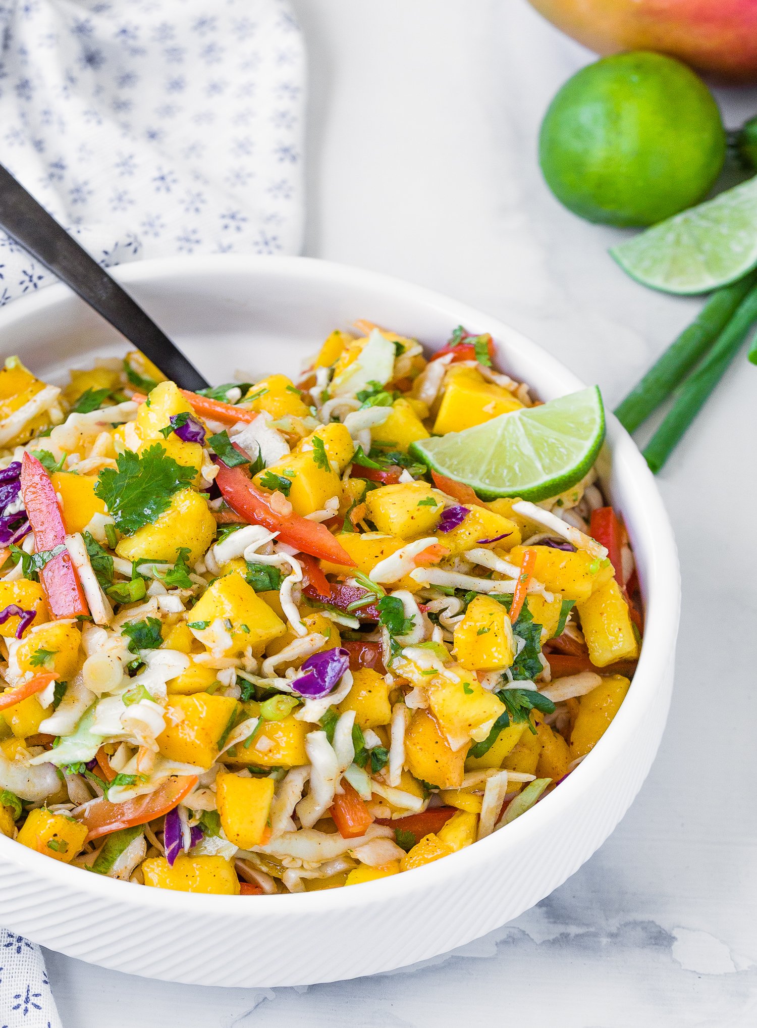 A bowl of mango slaw with red peppers, cabbage, and herbs, garnished with lime wedges.
