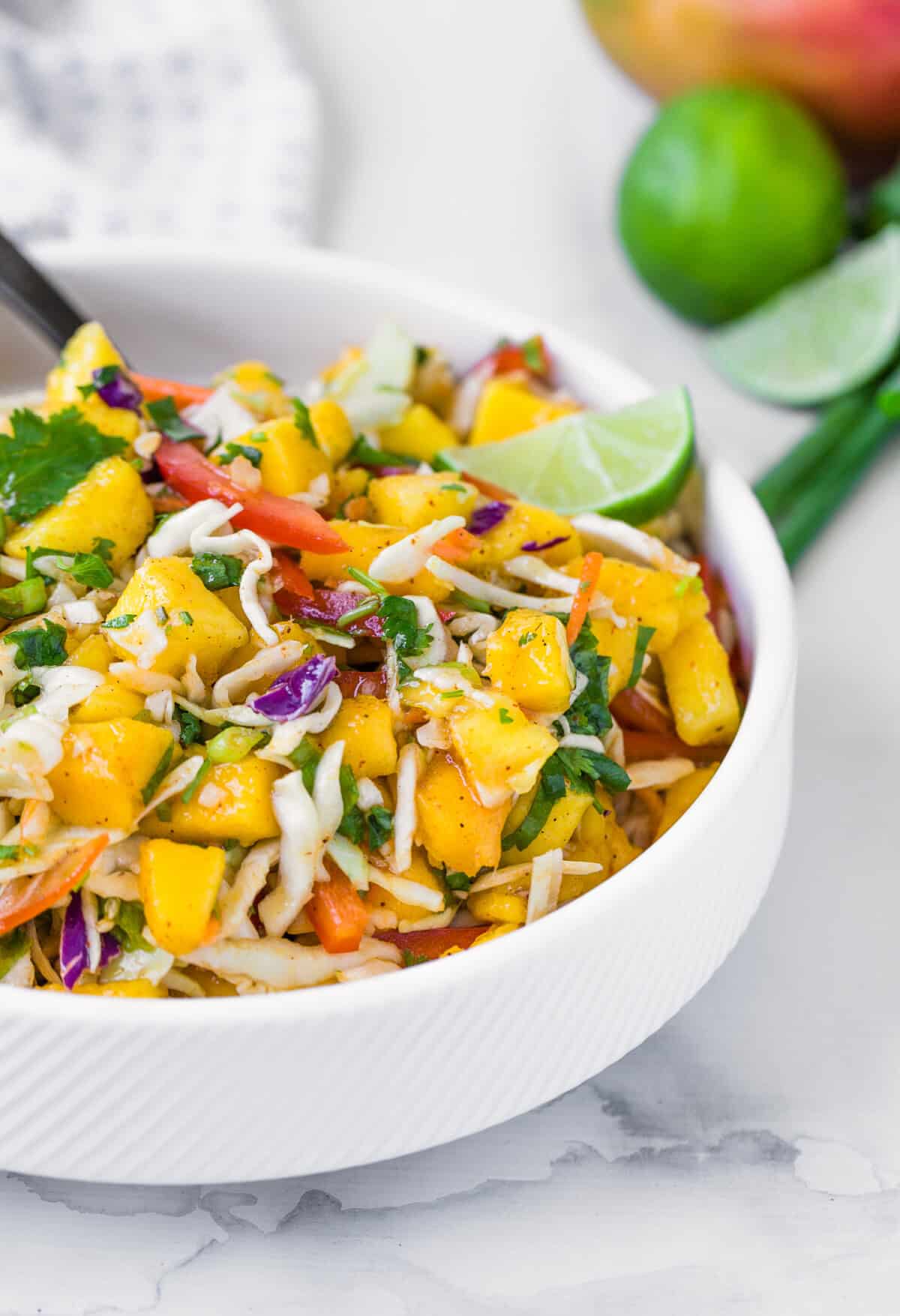 A fresh mango slaw in a white bowl with chopped vegetables, herbs, and lime wedges.