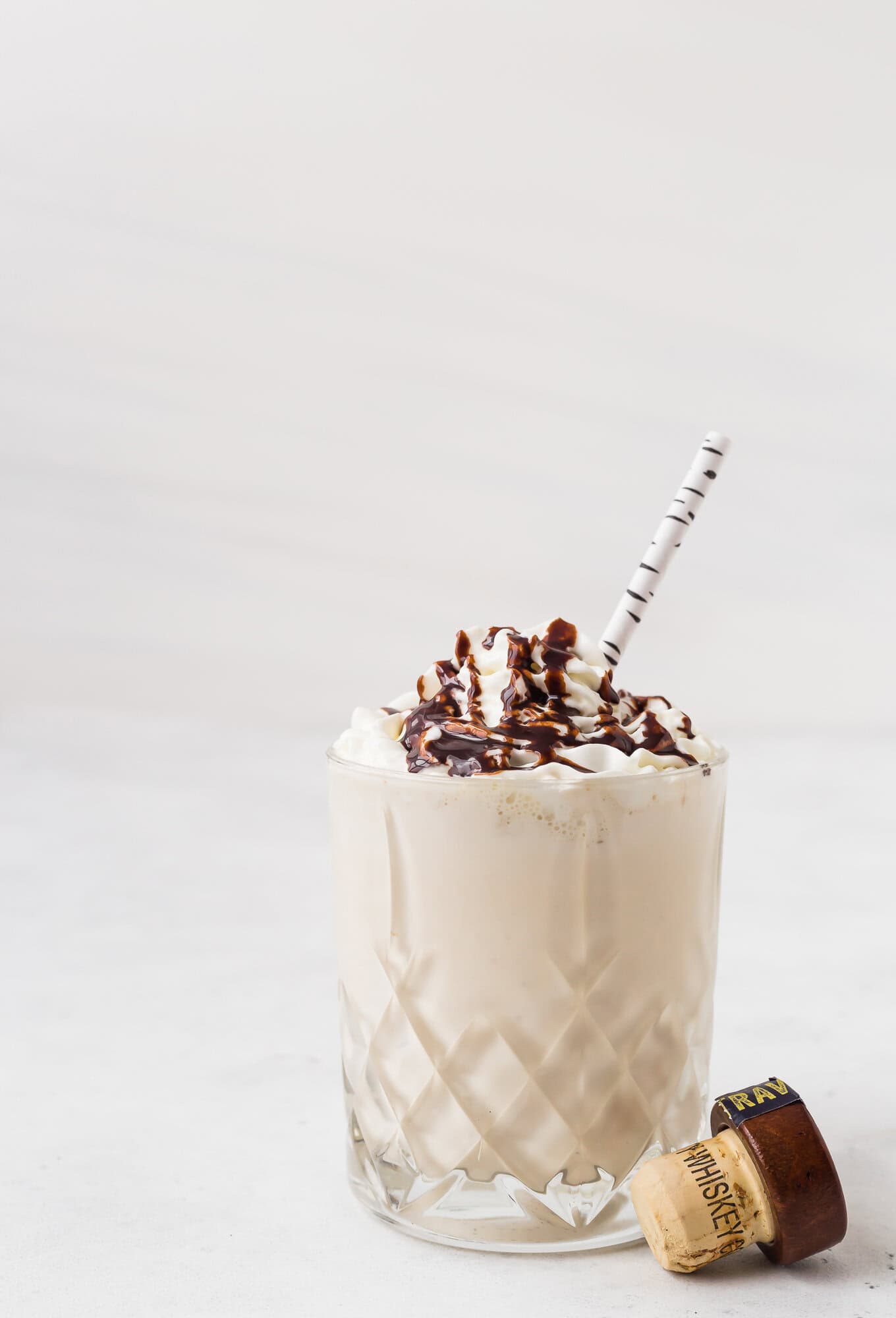 A glass of creamy milkshake topped with whipped cream and chocolate drizzle, with a striped straw and a bottle cap beside the glass.