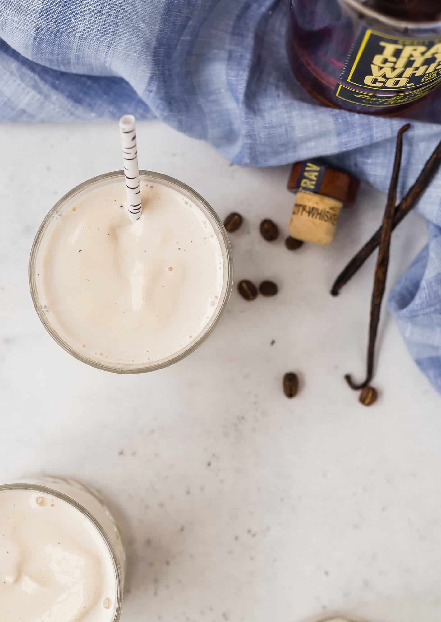 A creamy milkshake with a straw sits on a white surface near coffee beans, a cork, vanilla beans, and a bottle, with a blue cloth partially visible.