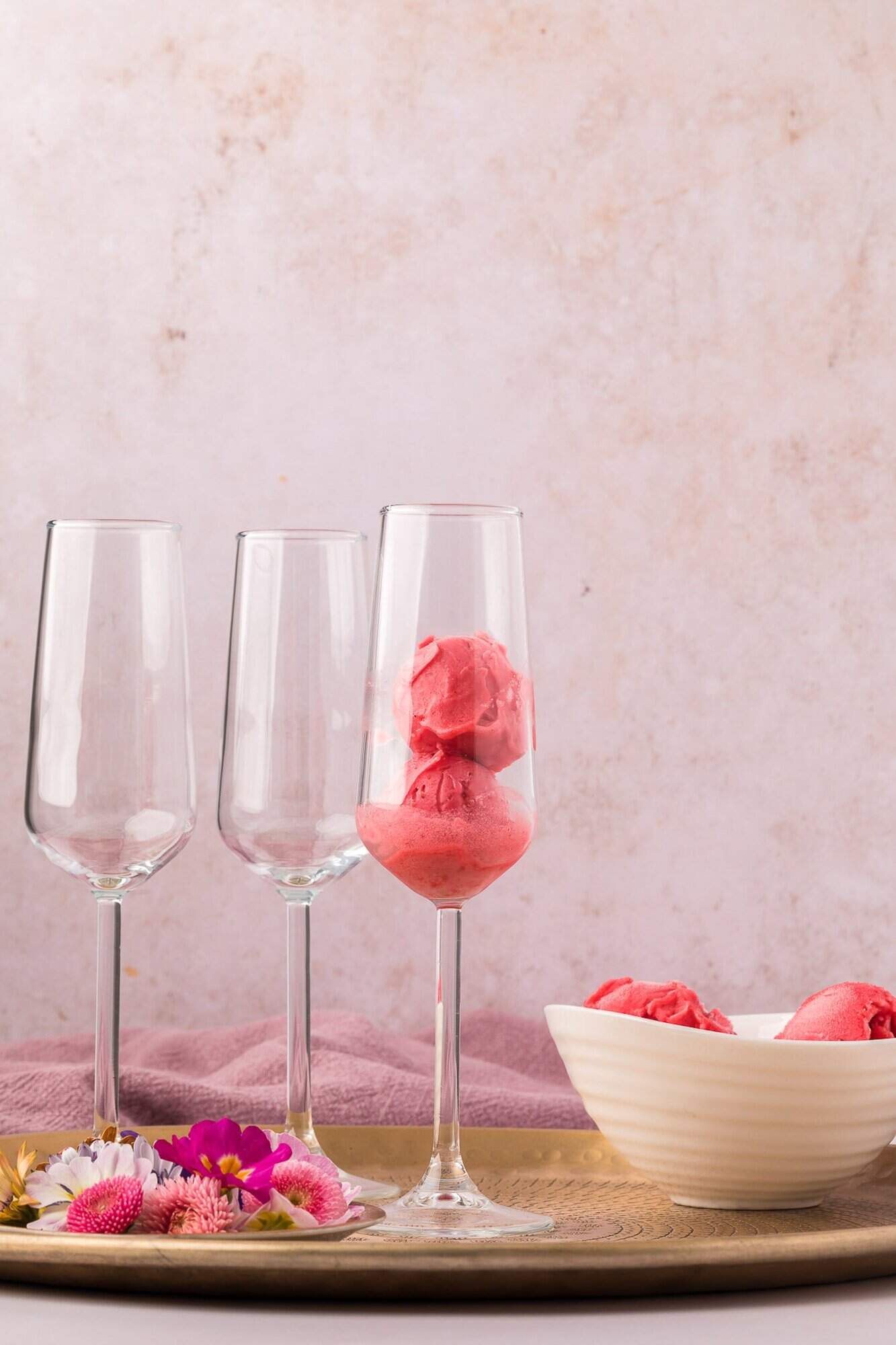 Raspberry Sorbet Champagne Float 6 Three champagne flutes on a tray; one contains scoops of pink sorbet. A white bowl with more sorbet and some flowers are also on the tray, against a light pink background.