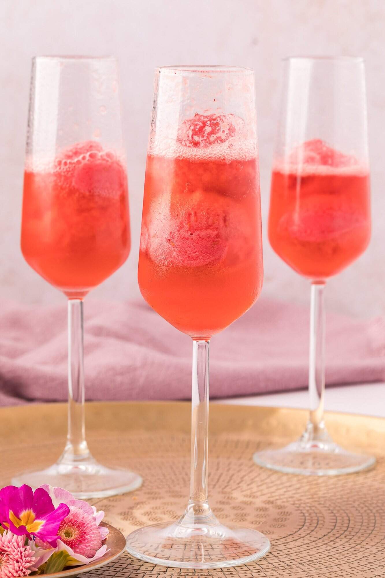 Raspberry Sorbet Champagne Float 10 Three champagne flutes filled with a pink fizzy drink and raspberry sorbet, placed on a tray with pink flowers and a mauve cloth in the background.