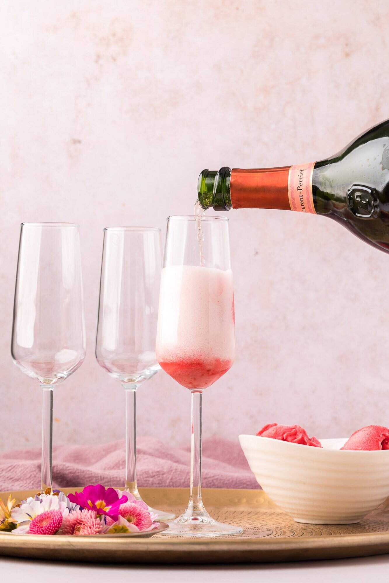 Raspberry Sorbet Champagne Float 8 A bottle pours sparkling wine into a glass with pink sorbet; two empty glasses, flowers, and a bowl of sorbet are on a tray nearby.