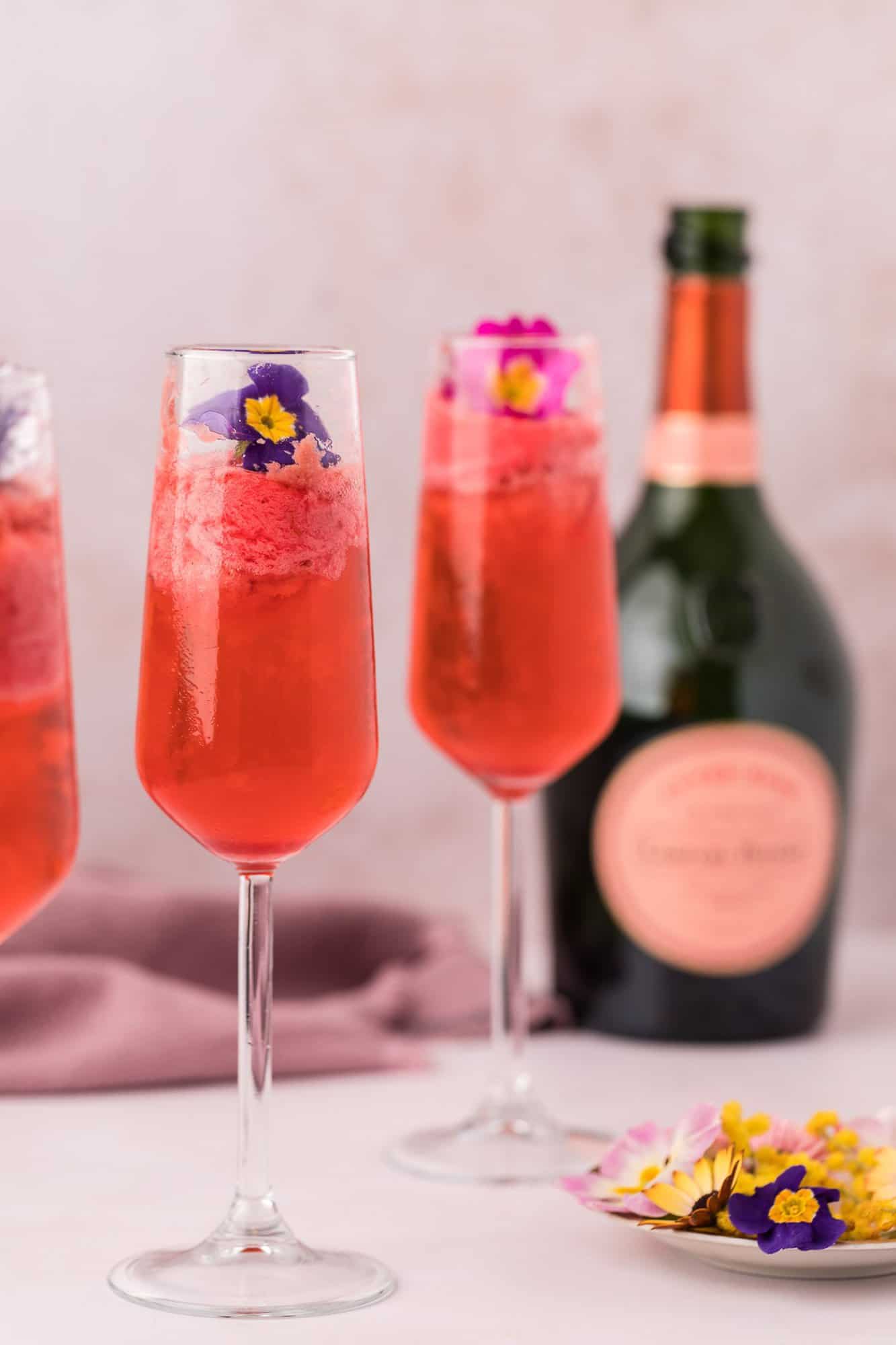 Raspberry Sorbet Champagne Float 2 Three champagne flutes filled with a pink champagne float and garnished with edible flowers, with a bottle of sparkling wine and a small plate of flowers in the background.