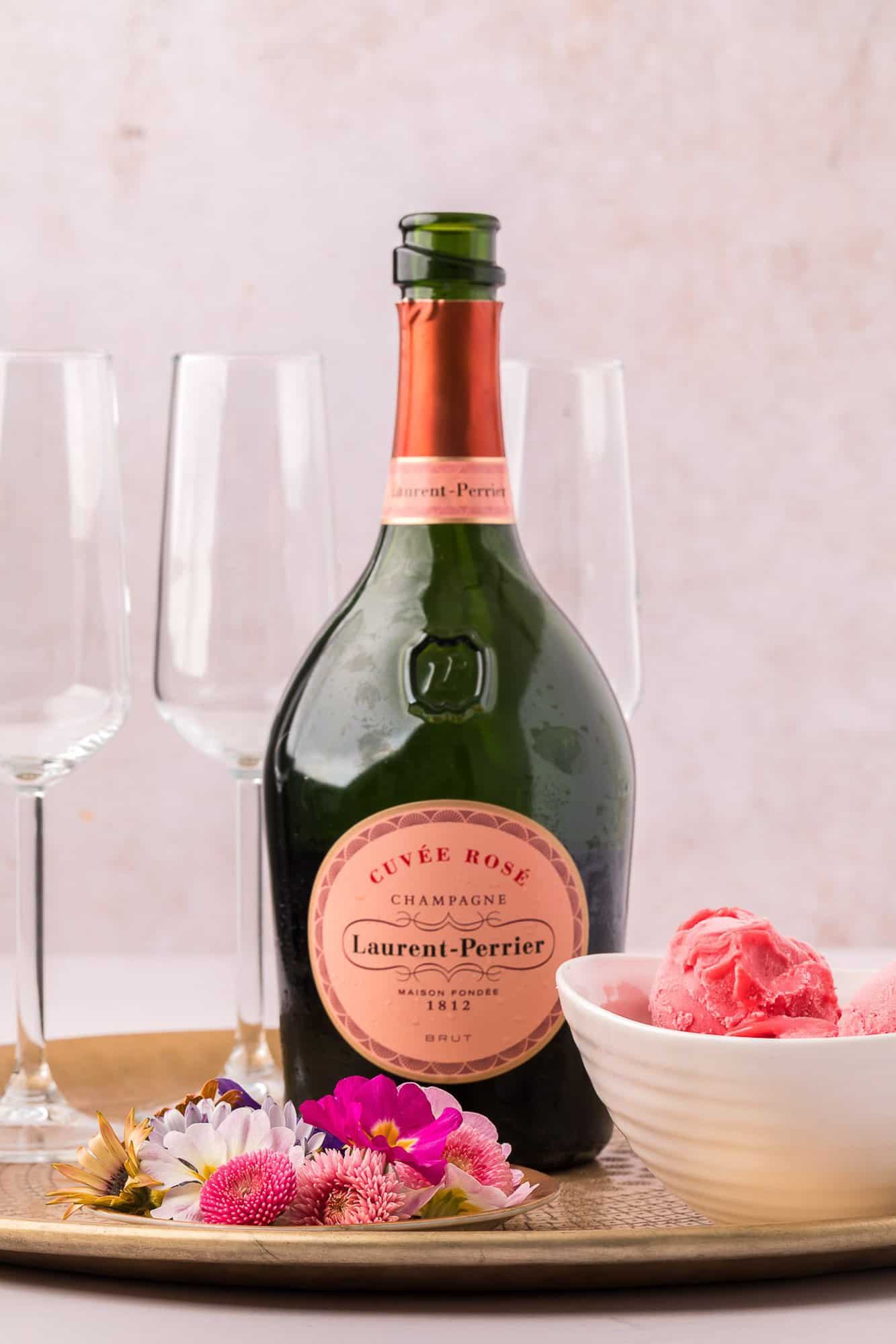 Raspberry Sorbet Champagne Float 4 A bottle of Laurent-Perrier Cuvée Rosé champagne on a tray with four empty champagne flutes, a bowl of pink sorbet, and some colorful flowers.