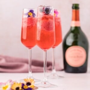 Three champagne flutes filled with a red cocktail, garnished with edible flowers, are placed beside an unopened bottle of sparkling wine on a light surface.