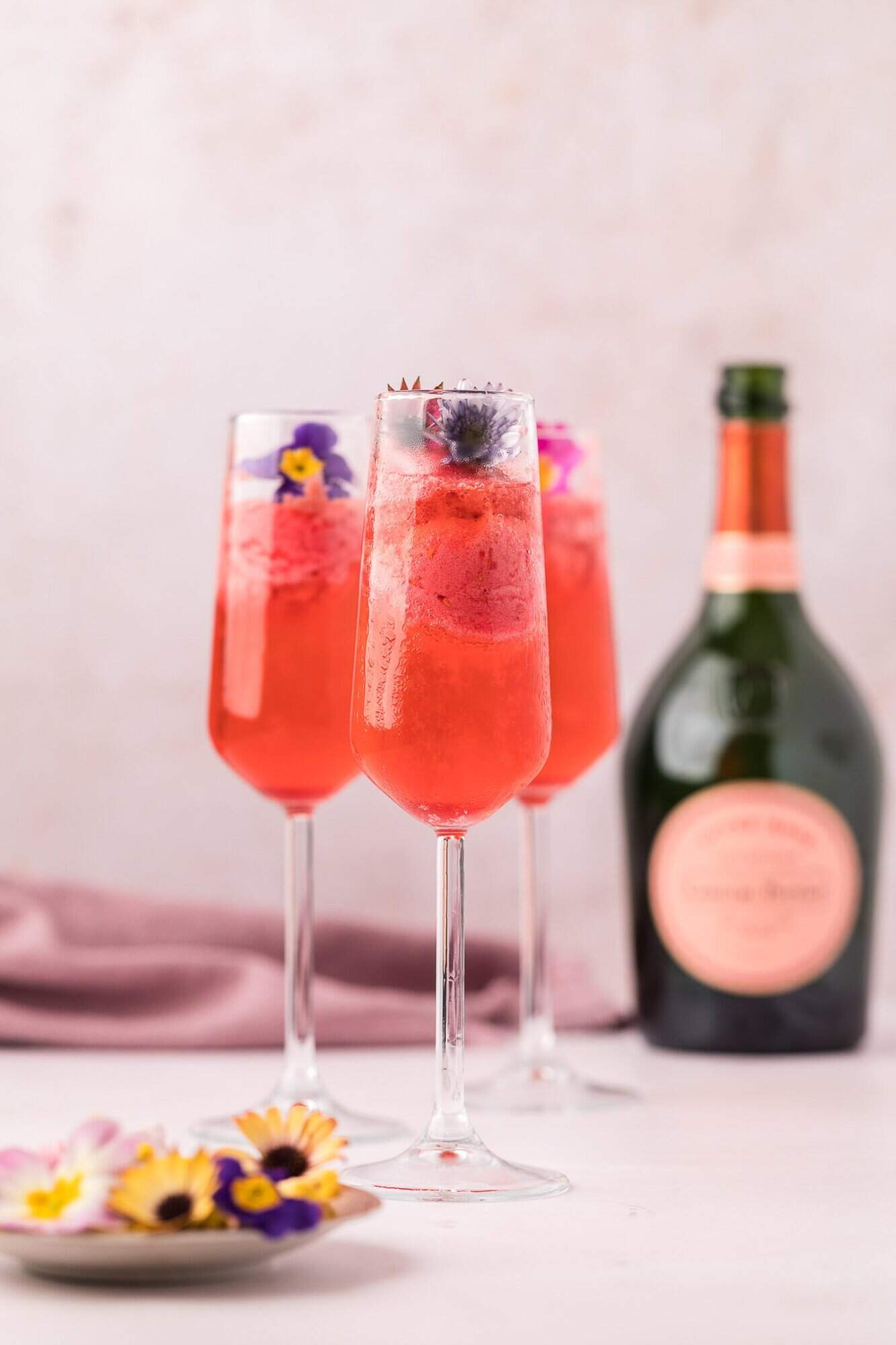 Raspberry Sorbet Champagne Float 12 Three champagne flutes filled with a pink cocktail garnished with edible flowers, with a bottle of sparkling wine in the background.