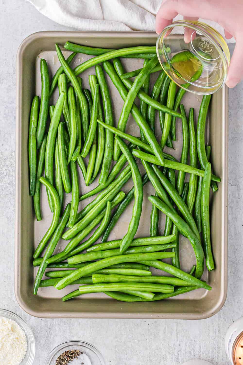 Person pouring olive oil over fresh green beans arranged on a baking tray, with small bowls of ingredients like pepper and cheese nearby.