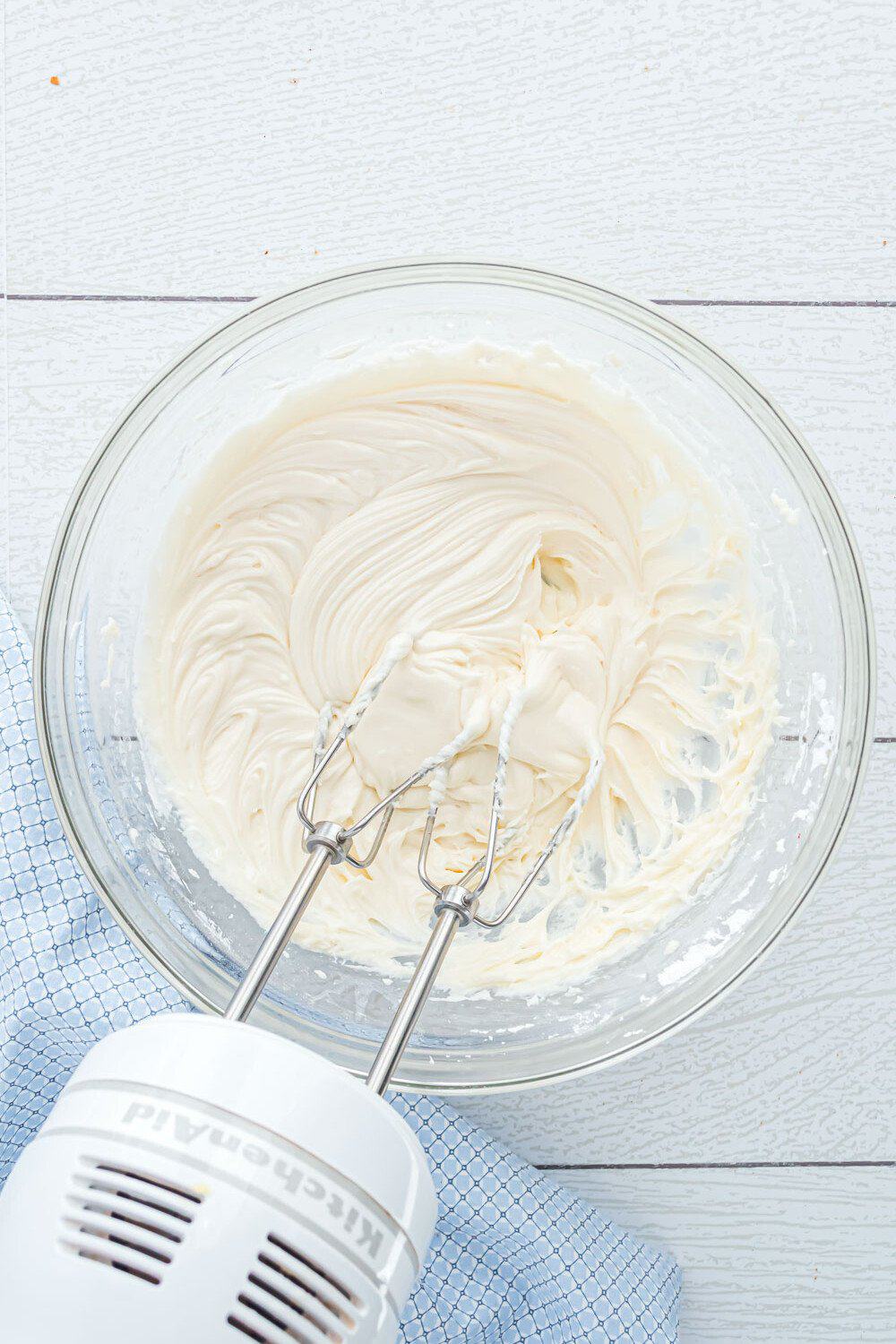 A hand mixer with beaters is resting in a glass bowl of creamy, whipped mixture on a light wooden surface with a blue cloth nearby.