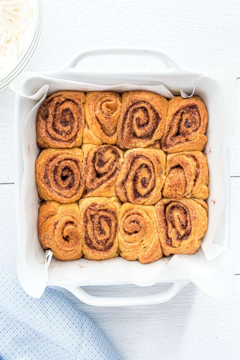 A white baking dish lined with parchment paper holds twelve baked cinnamon rolls arranged in a 3x4 grid.