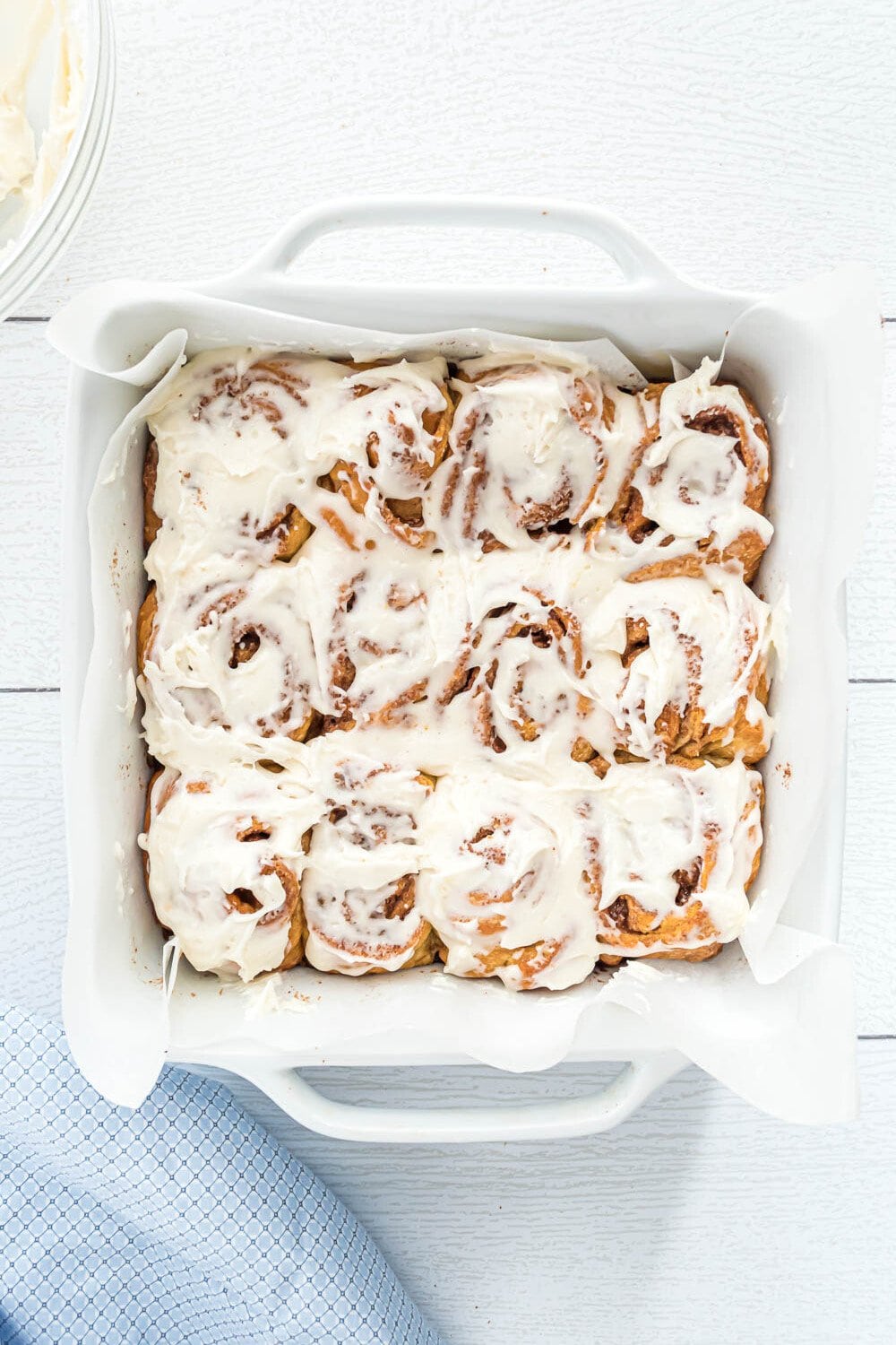 A white baking dish filled with twelve cinnamon rolls topped with white icing, placed on a white surface next to a blue cloth.