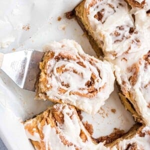 A close-up of a frosted cinnamon roll being lifted from a baking dish with a metal spatula.
