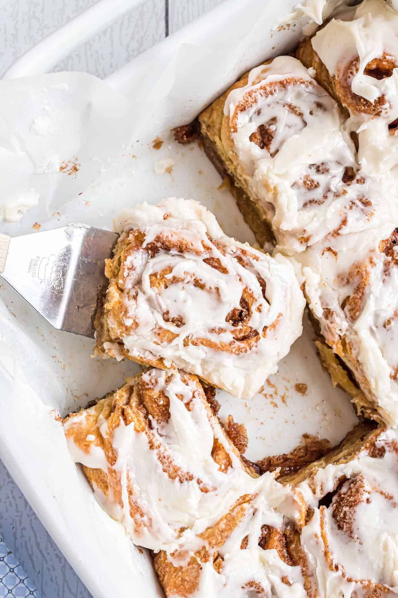 A spatula lifts a frosted cinnamon roll from a baking dish containing several iced cinnamon rolls.