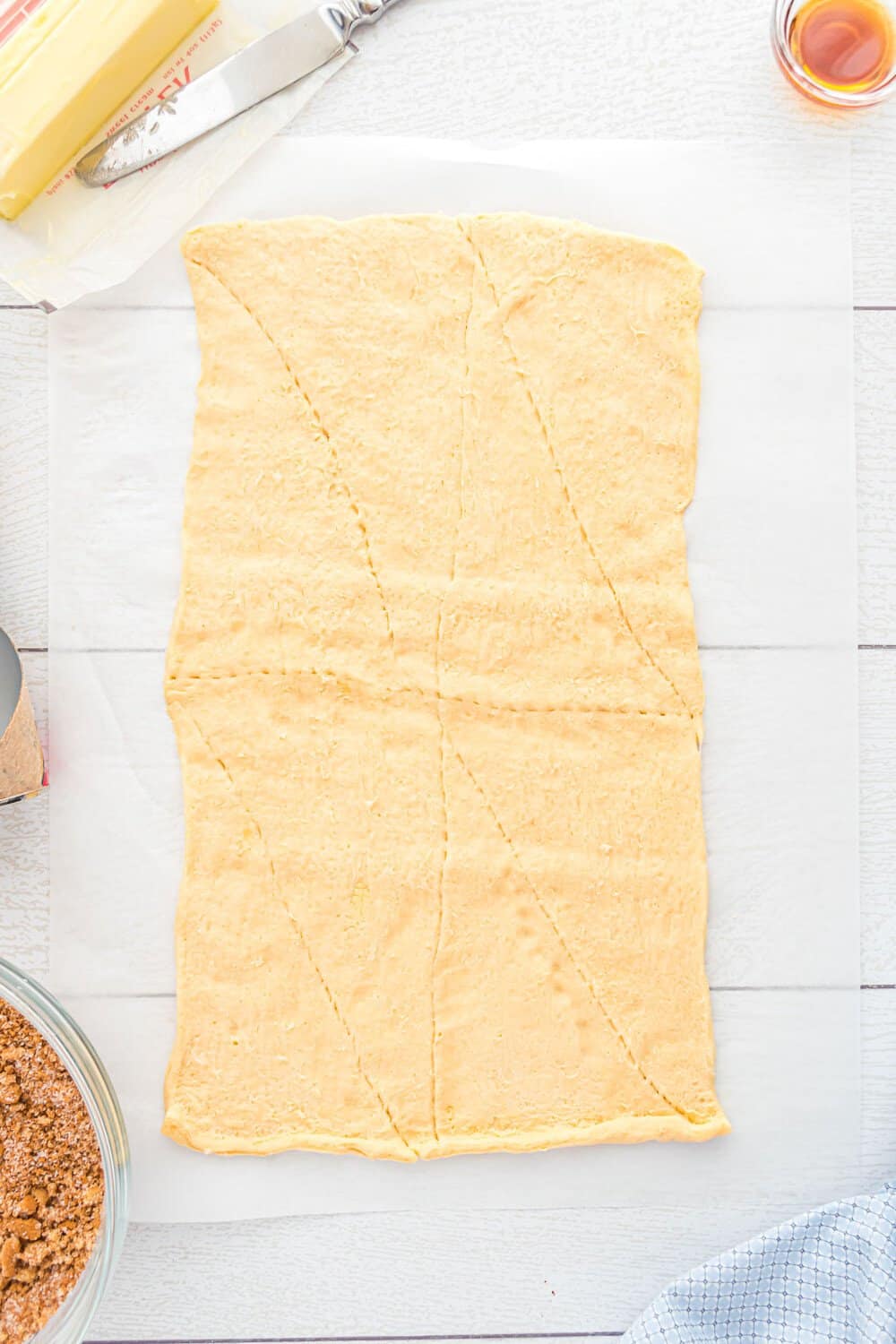 Rectangle of raw dough on parchment paper, scored into triangles. Butter, a bowl of brown sugar mixture, and a knife are nearby on a light-colored surface.