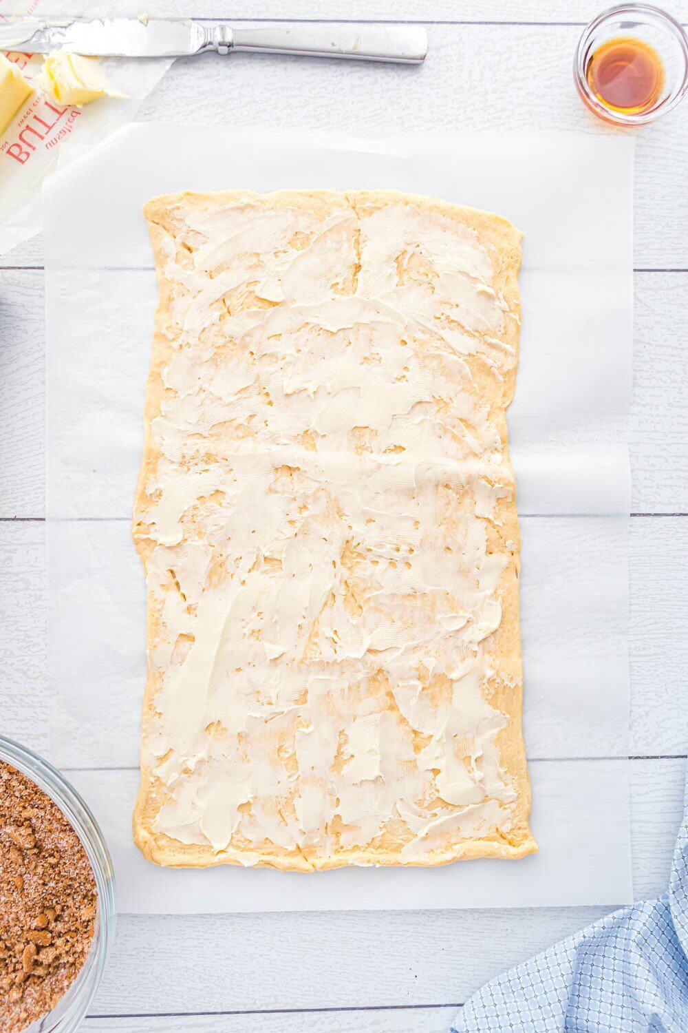 Rectangular sheet of dough spread with a layer of butter on parchment paper, surrounded by a bowl of crumb mixture, butter, a knife, and a small cup of liquid.