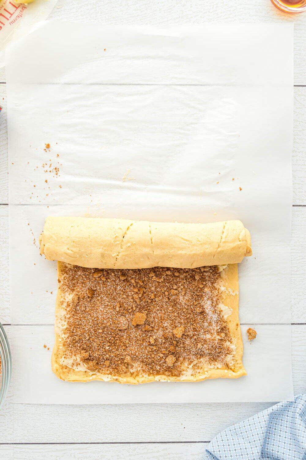 A rolled sheet of dough sprinkled with cinnamon sugar is being rolled up on parchment paper on a light wooden surface.