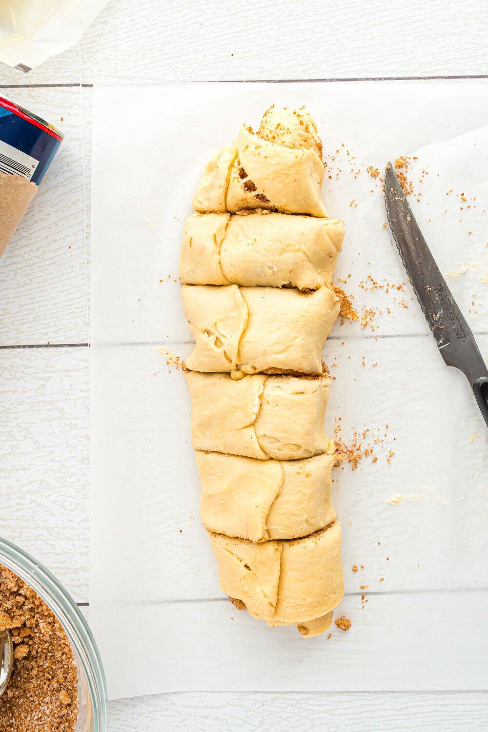Dough log sliced into pieces on parchment paper, with a serrated knife and crumbs nearby. A bowl and packaging are partially visible at the edges.