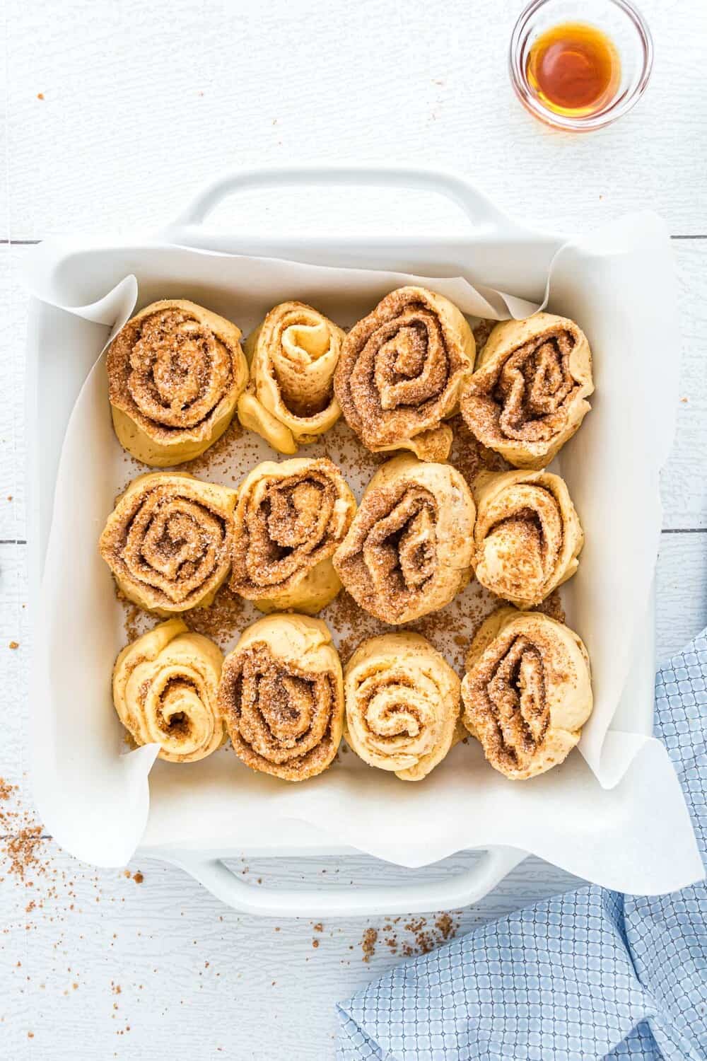 A square baking dish lined with parchment paper holds twelve unbaked cinnamon rolls. A small glass of vanilla extract and a blue cloth are nearby on a white wooden surface.