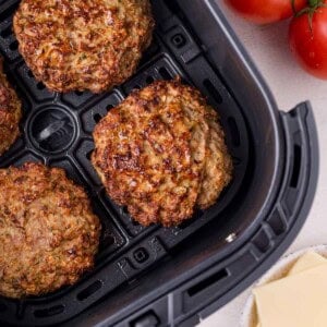 Four cooked burger patties in an air fryer basket, with fresh tomatoes and slices of cheese visible beside the basket.