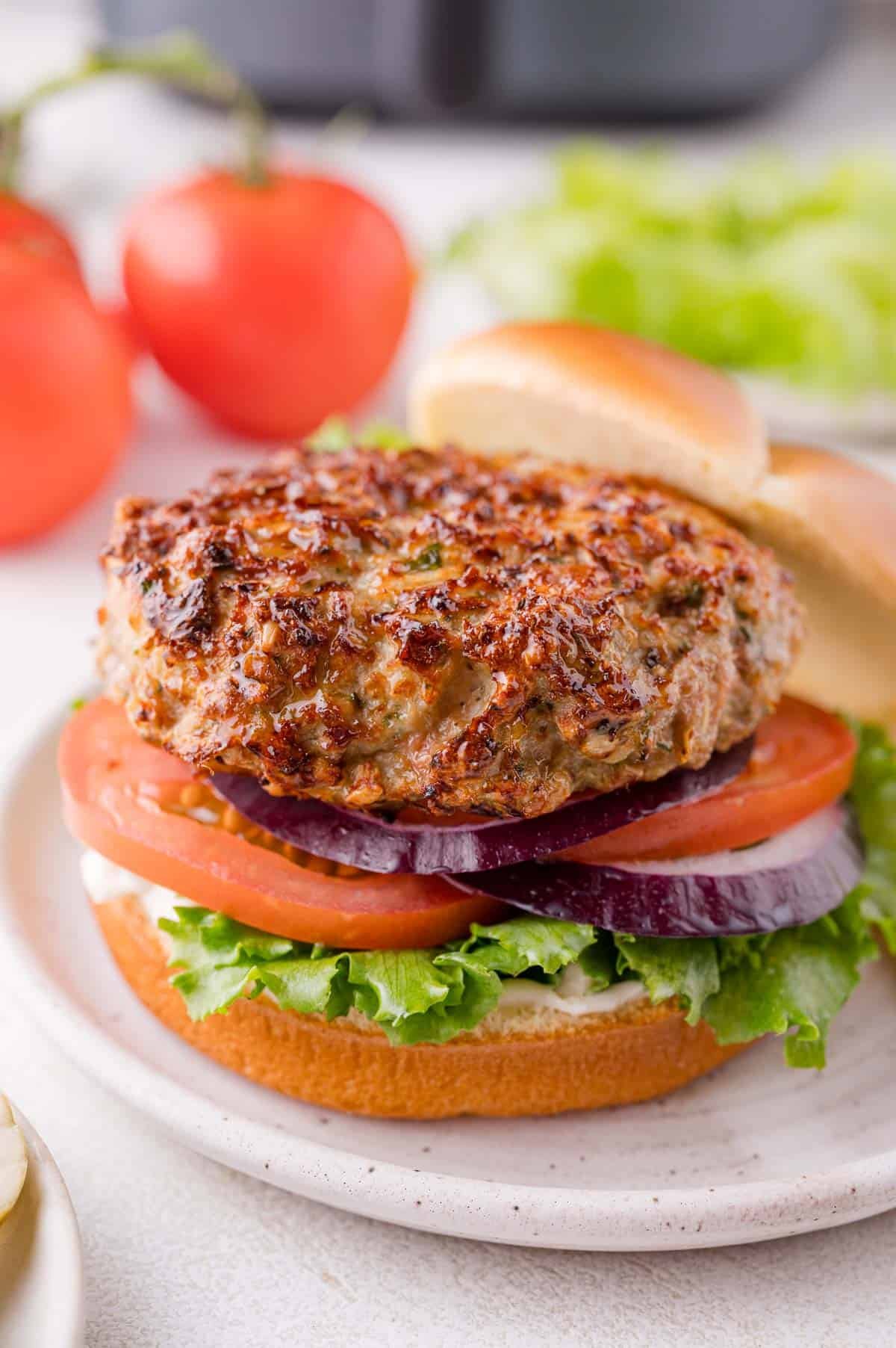 A chicken burger with lettuce, tomato, and red onion on a bun, served on a plate with tomatoes and lettuce in the background.