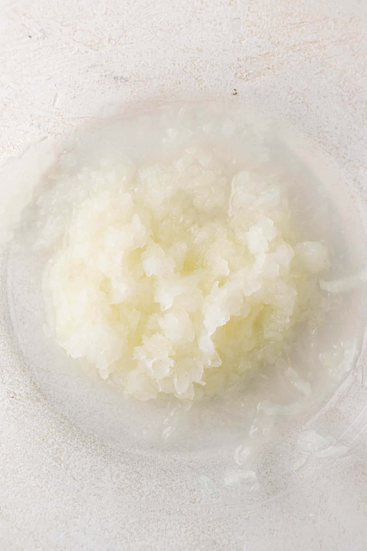 Finely grated or minced white onion with juice collected in a clear glass bowl on a light surface.