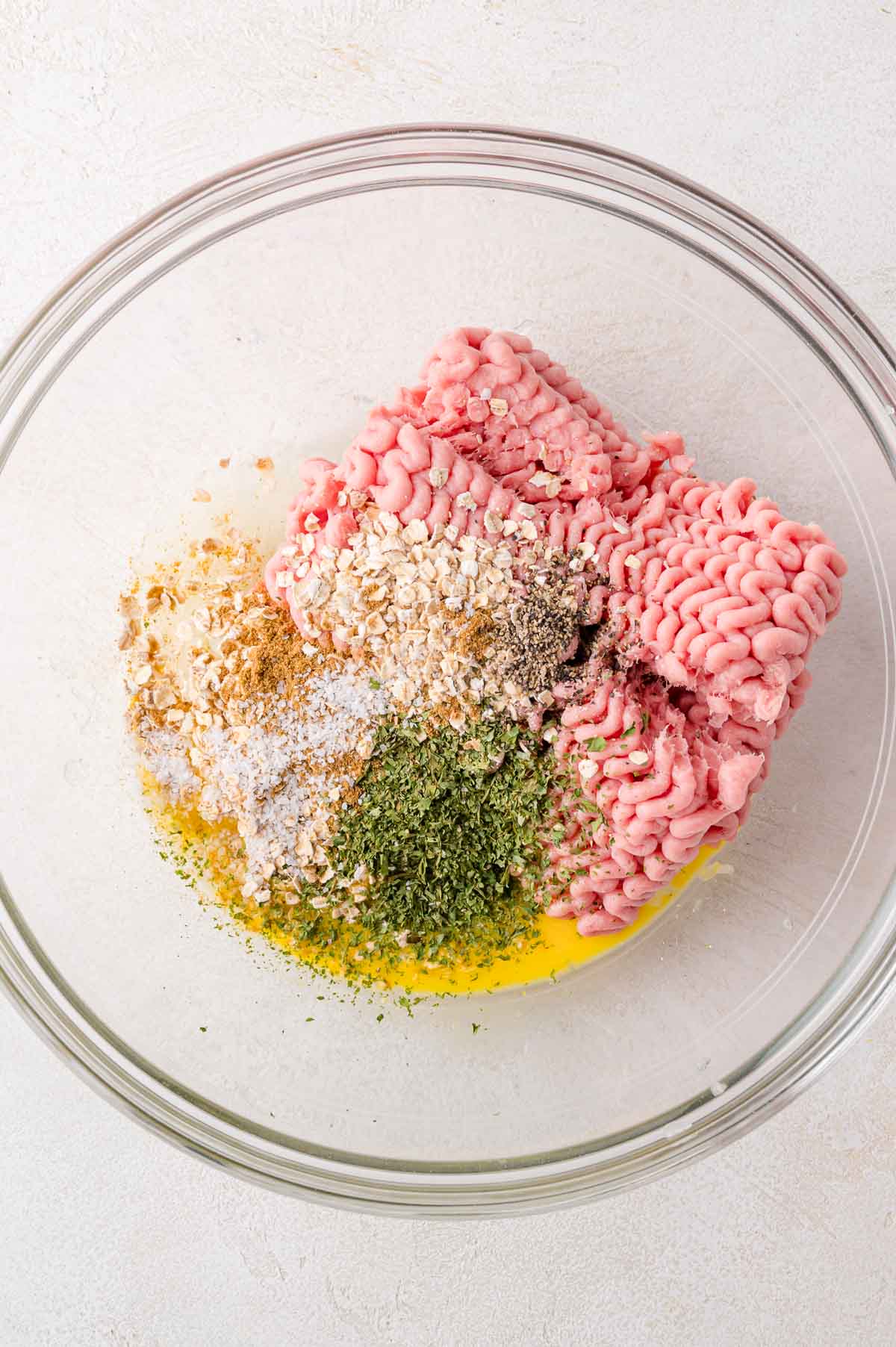 Ground meat, egg, oats, dried herbs, salt, pepper, and spices are arranged in a large clear glass mixing bowl on a white surface.