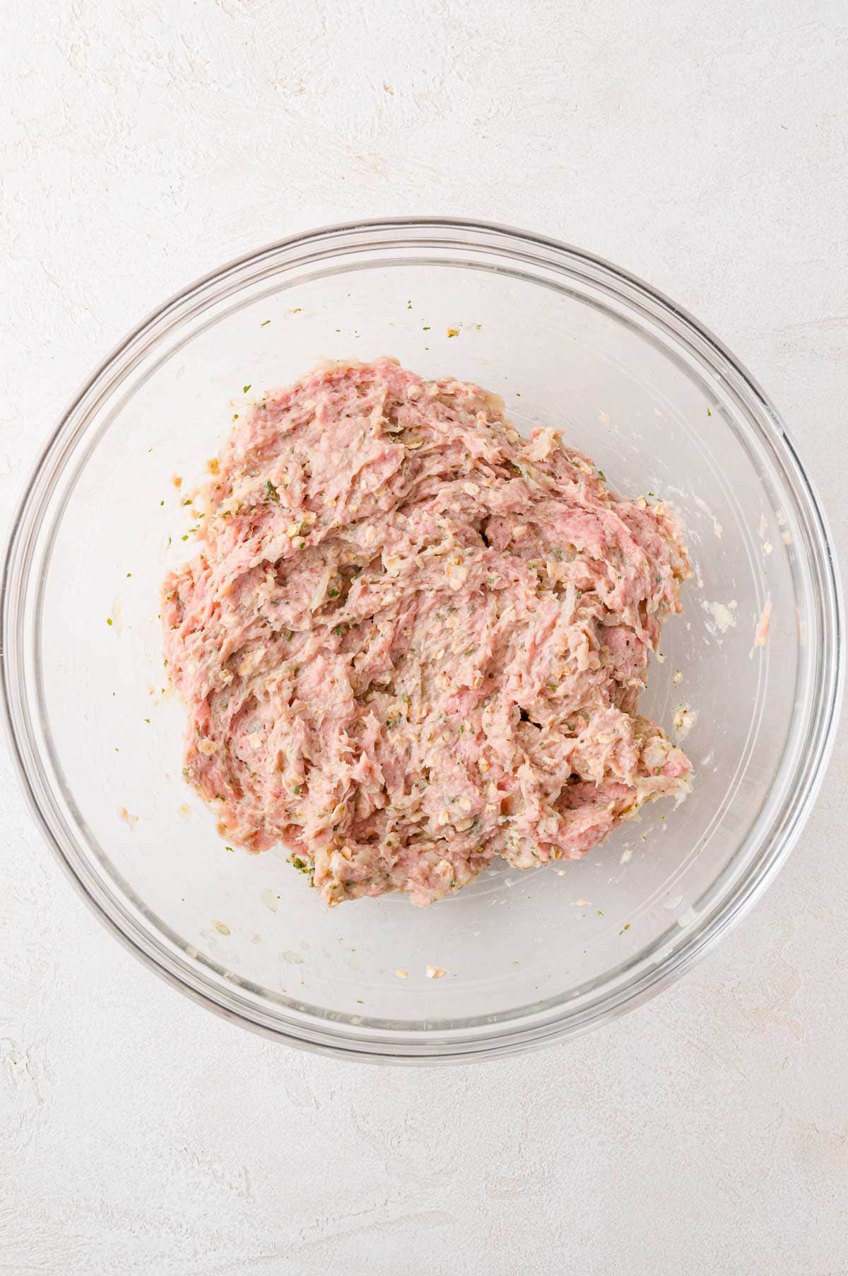 Ground meat mixture with herbs and seasonings in a clear glass bowl, placed on a light-colored surface.