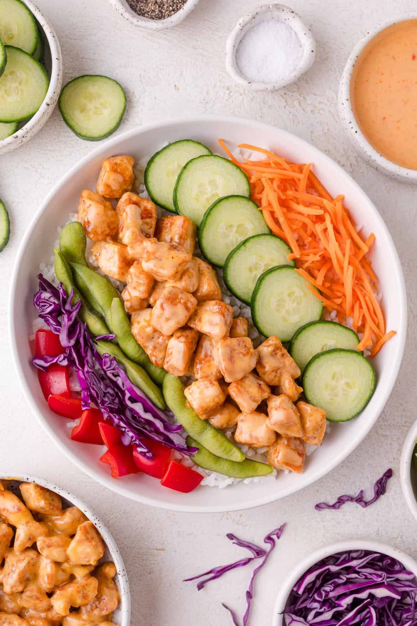 A bowl of rice topped with bang bang chicken, sliced cucumber, shredded carrots, red bell pepper, edamame, and purple cabbage, surrounded by small bowls of sauce and vegetables.